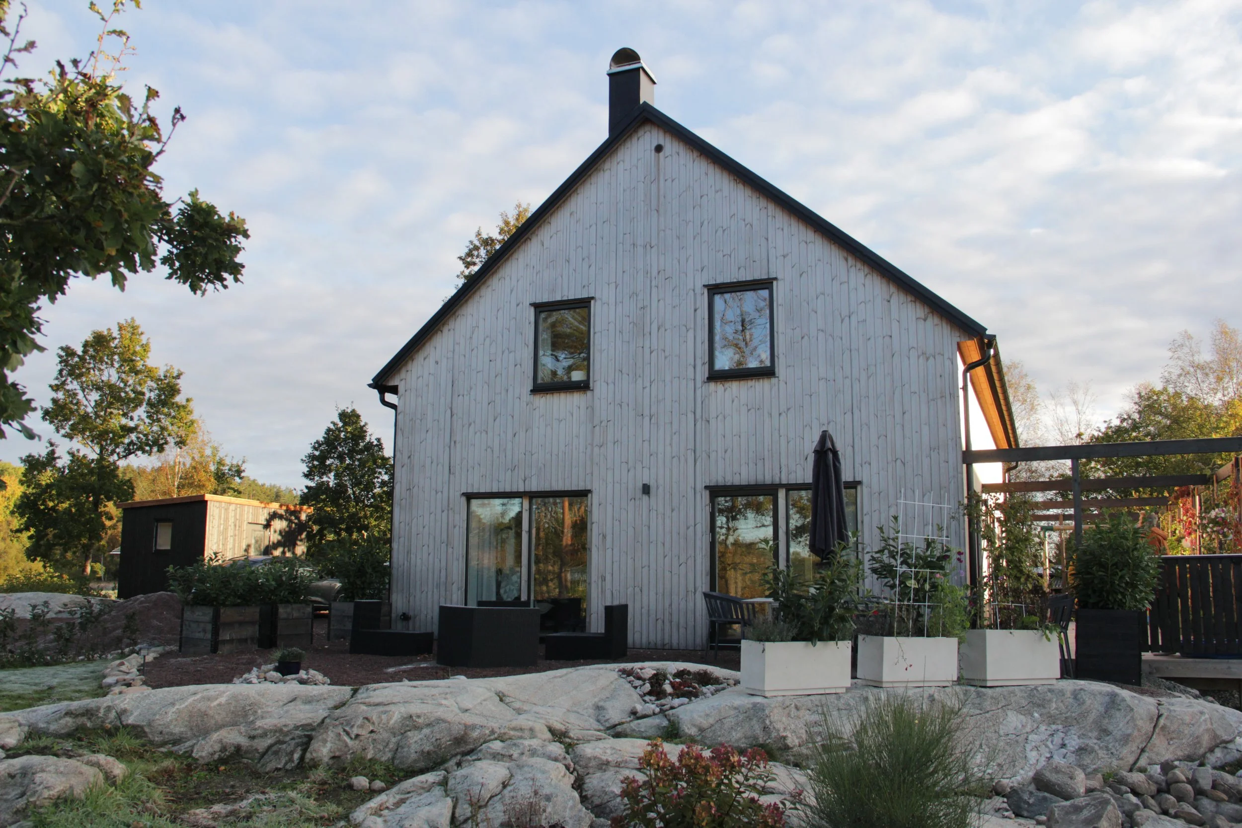 Modernt hus med träfasad och stora fönster, omgiven av träd och stenar i en trädgård.