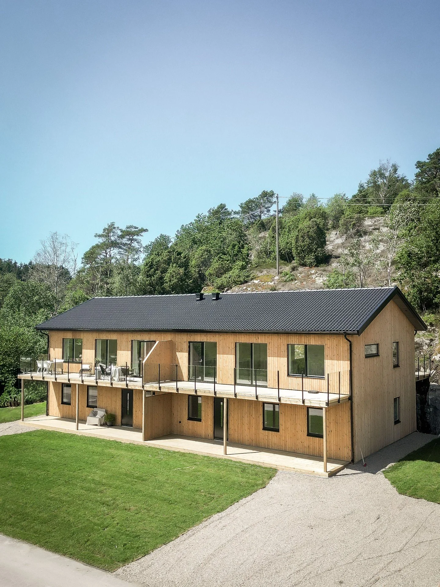 Tvåvåningshus i trä med svart tak, balkonger och grön gräsmatta framför, omgiven av träd och en klippa i bakgrunden.