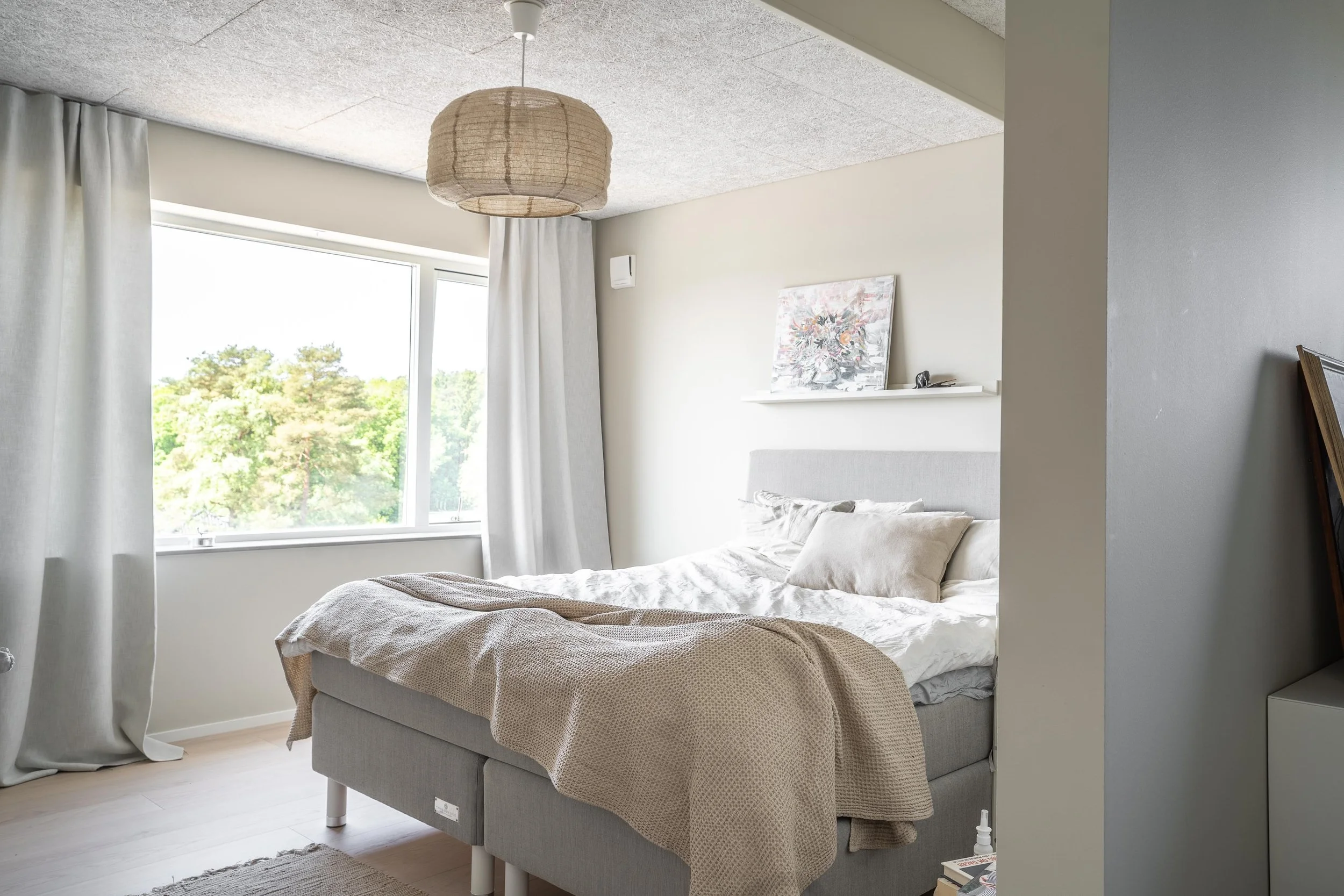 Ett ljust och modernt sovrum med en stor säng täckt av kuddar och en beige filt. Stora fönster med vita gardiner släpper in naturligt ljus och en tavla finns på en hylla ovanför sängen. En hängande lampa i rotting hänger från taket. O Lind AB.
