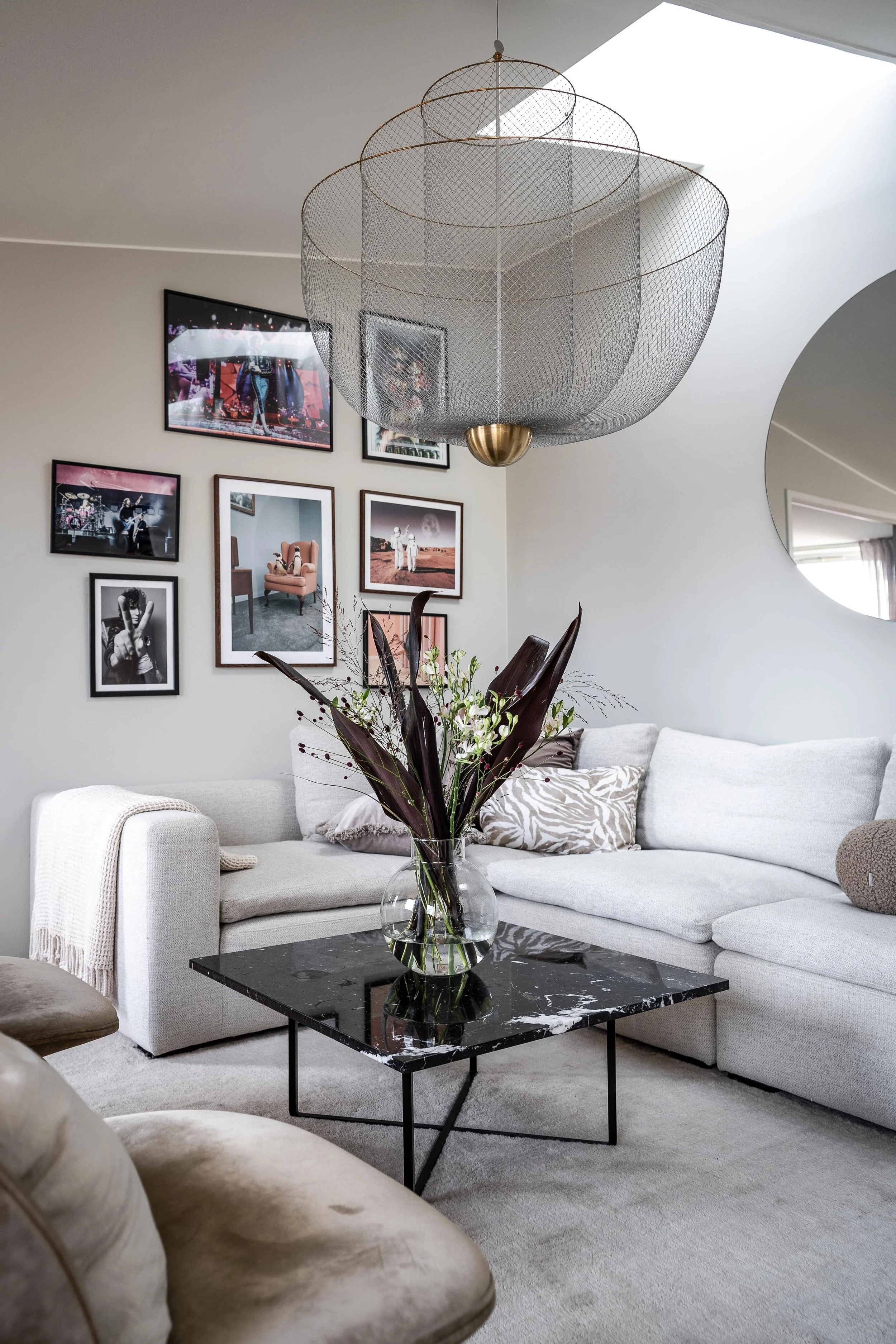 Modernt vardagsrum med ljusgrå hörnsoffa, svart marmorbord, och väggkonst. Stort dekorativt taklampa och rund spegel. Glasvas med blommor på bordet. O Lind AB.