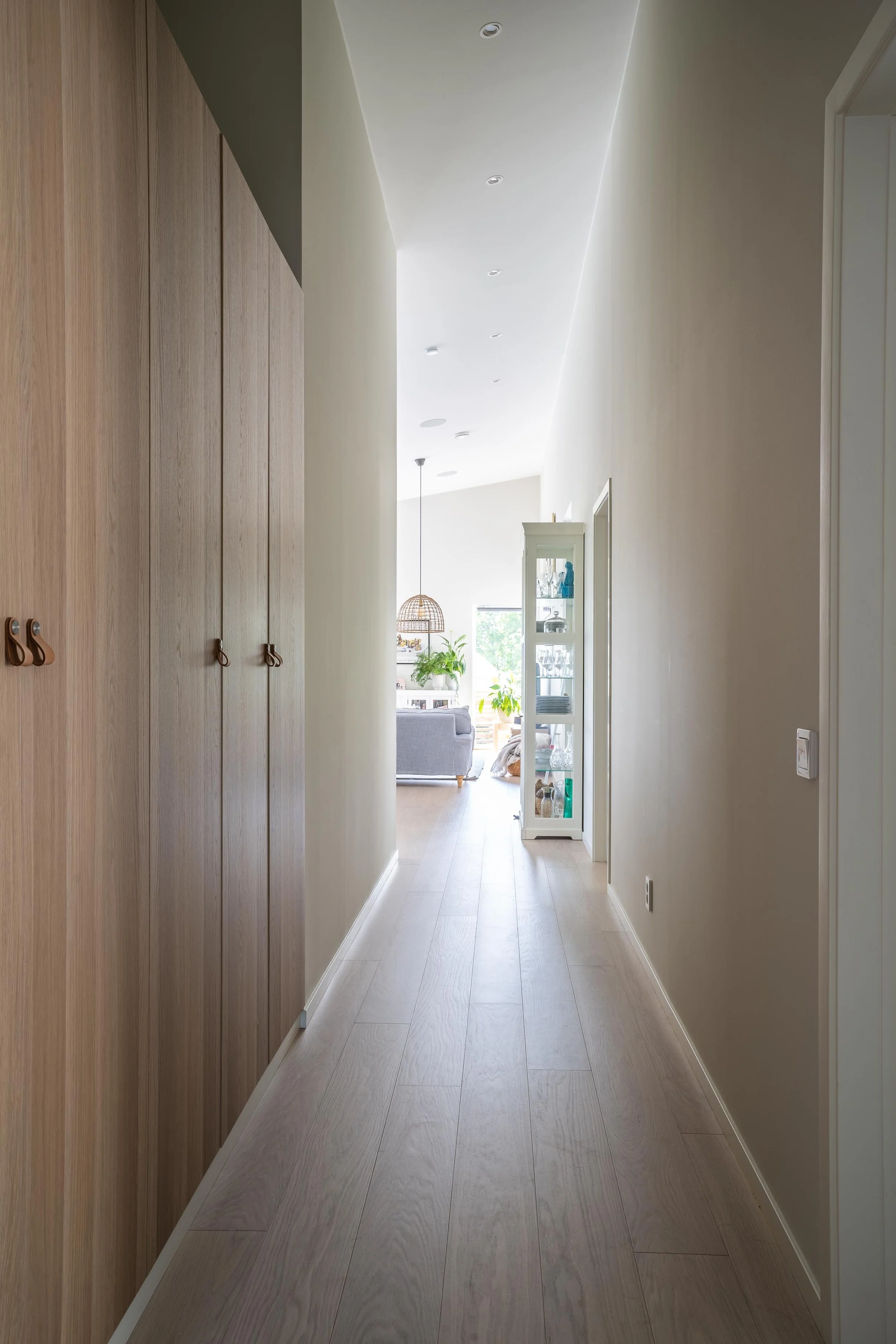 Smal hall med trägolv och ljusa väggar, garderob på vänster sida, vitrinskåp till höger, leder till ljust rum med stor fönster. O Lind AB.