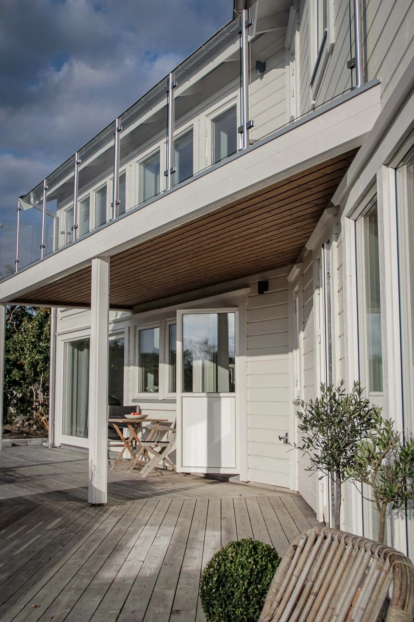 Vit modern altan med glasräcke och trädäck. Trädgårdsmöbler och gröna växter syns på terrassen.
