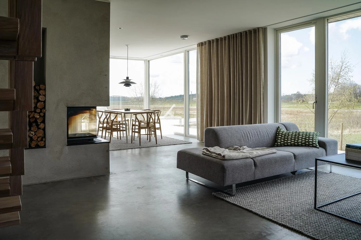 Modernt vardagsrum med öppen spis, grå soffa, glasbord, stora fönster, matbord med stolar och landskapsvy utanför. O Lind AB.