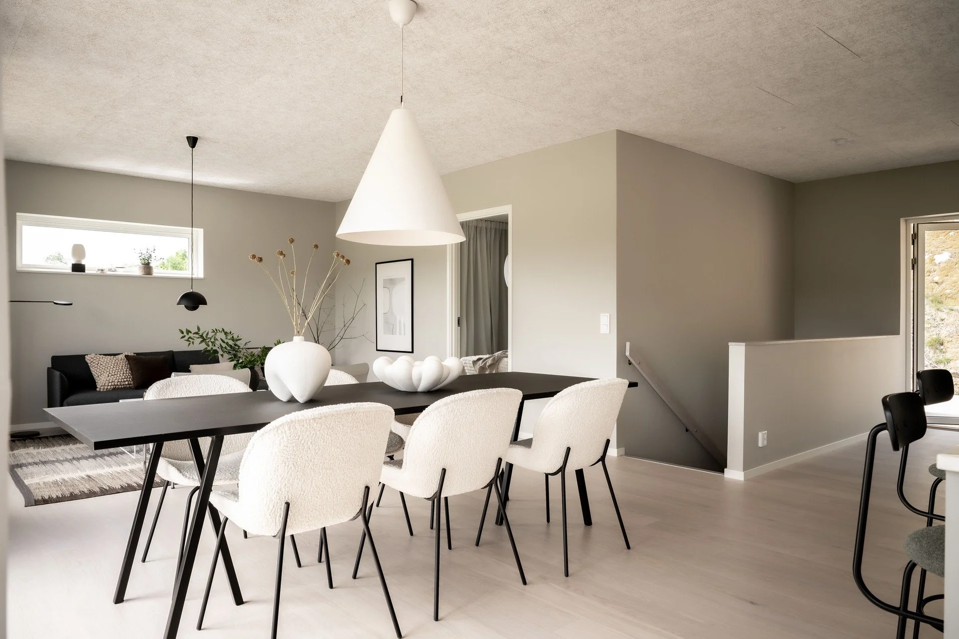 Modern matplats med ljusa väggar, svart bord, vita fåtöljer, och dekorativa vaser. I bakgrunden syns ett vardagsrum med svart soffa och växt. O Lind AB.