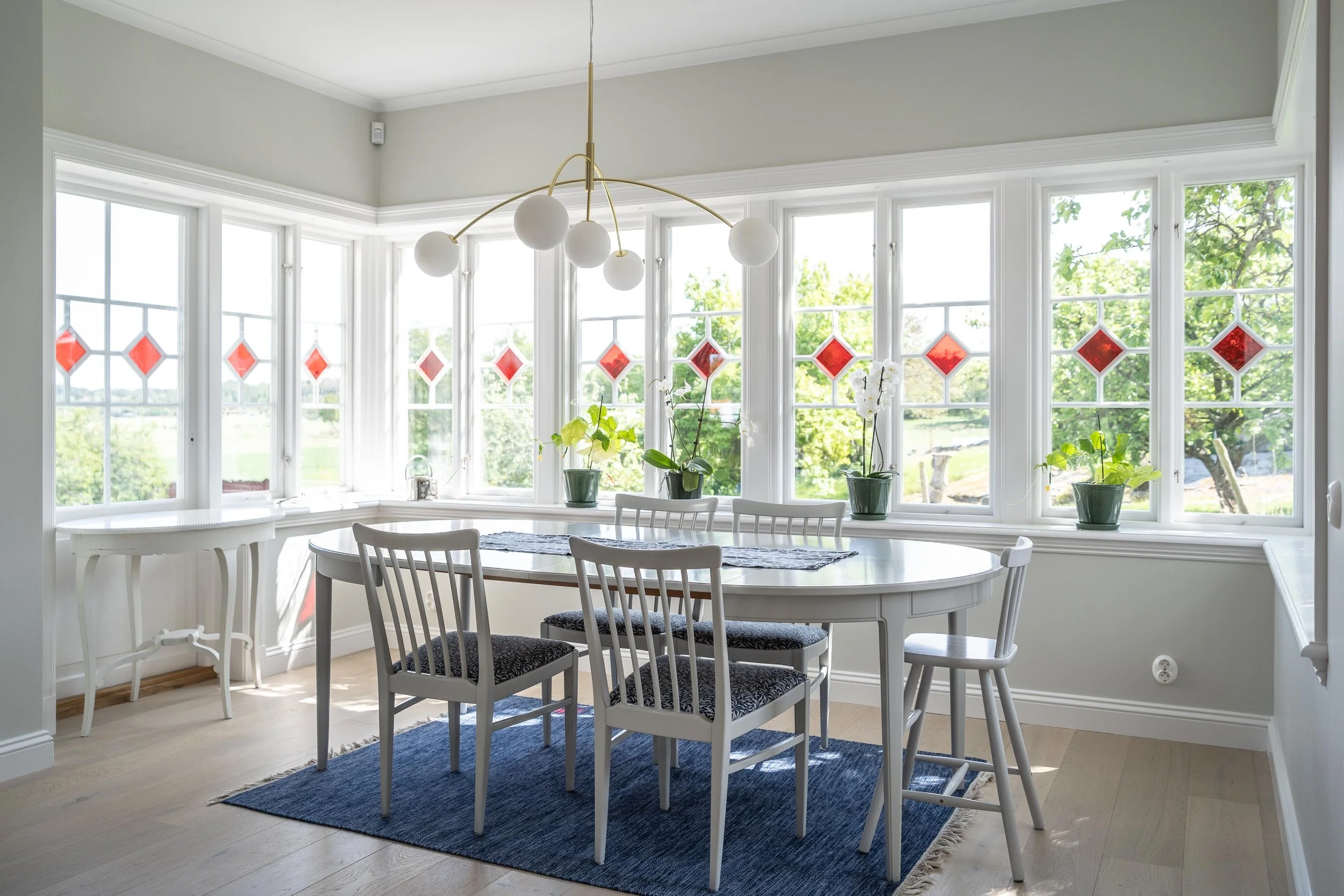 Matplats med vita stolar och bord, stora fönster med röda glasmönster, flera krukväxter och blå matta, modern ljuskrona i taket. O Lind AB.