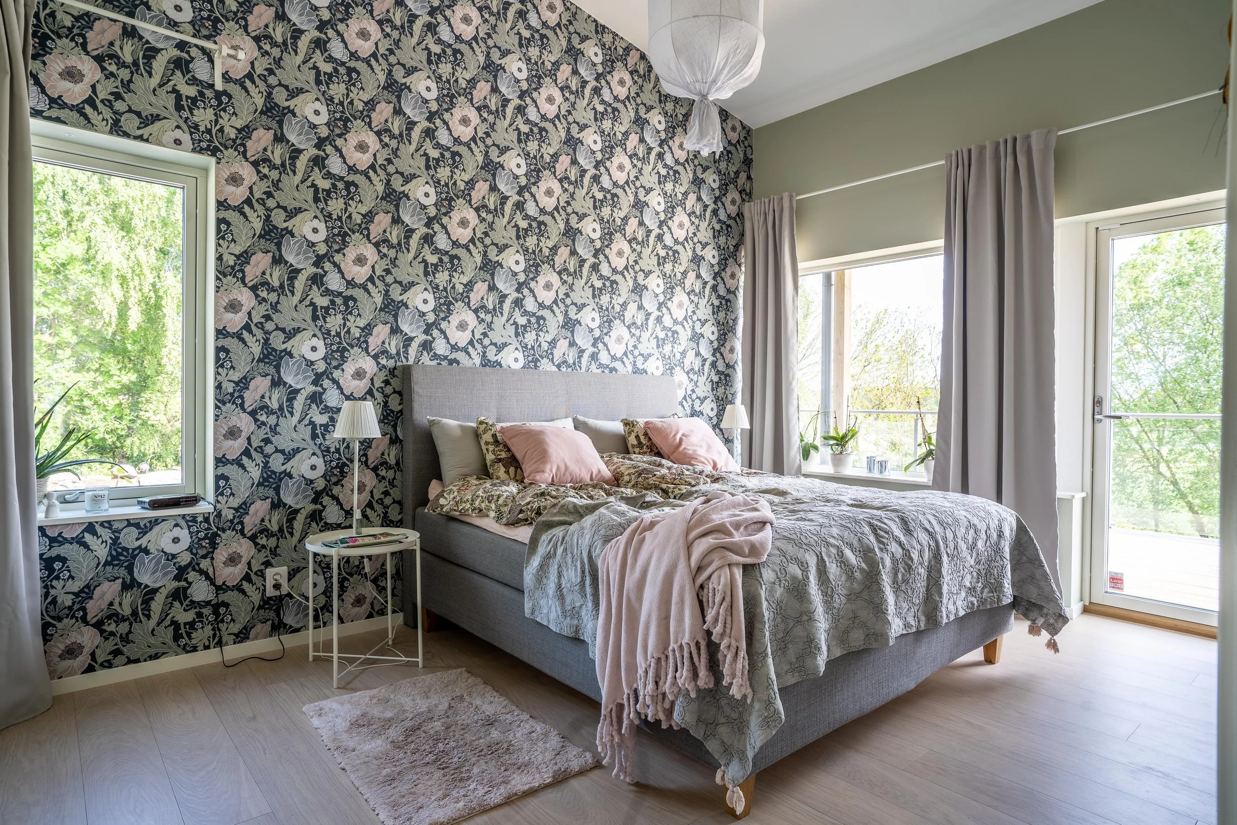 Sovrum med dubbelsäng, blommig tapet och stora fönster; gråa gardiner, rosa kuddar och filt. O Lind AB.