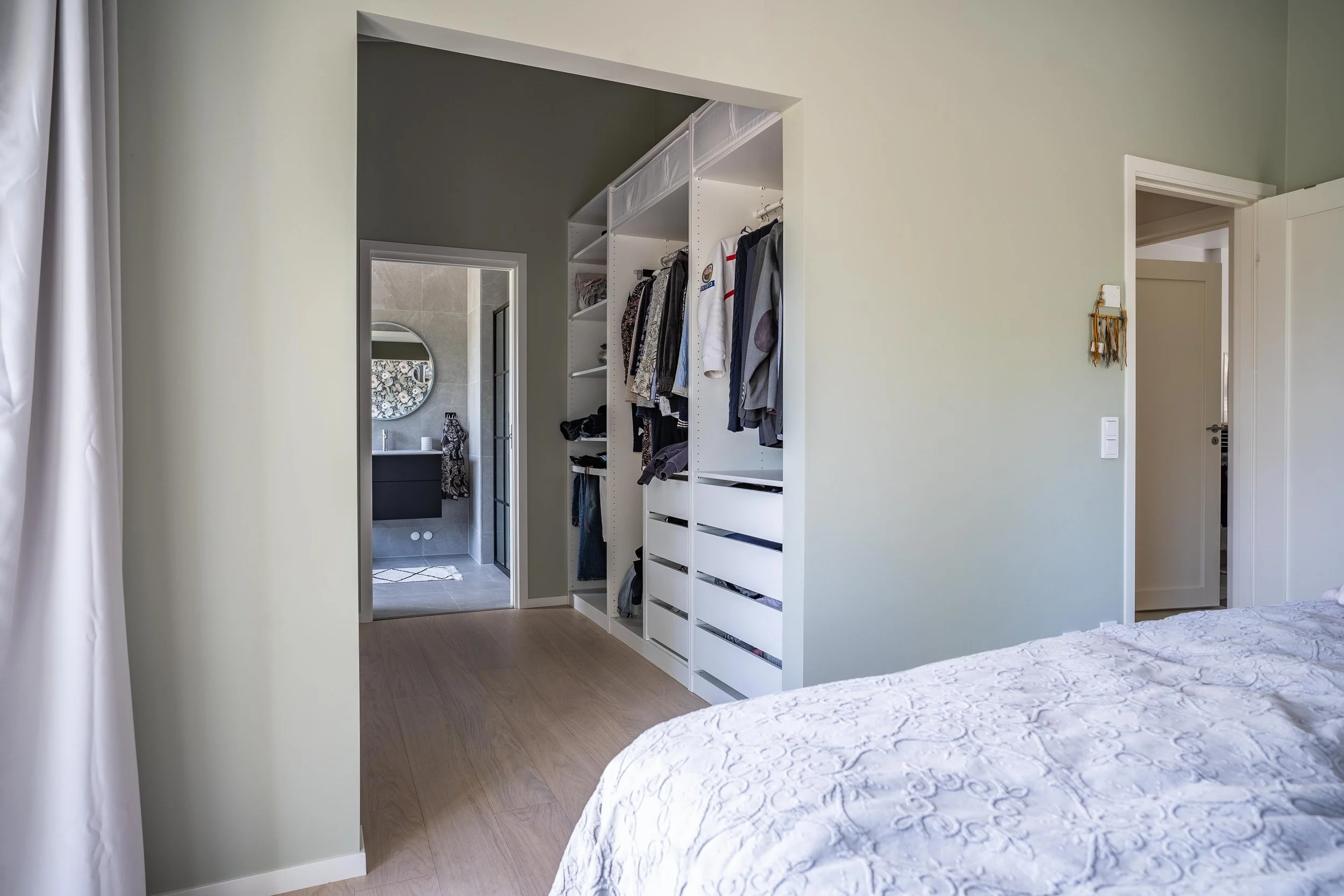 Sovrum med klädkammare och badrum i bakgrunden, vita väggar och dörrar, hängande och vikta kläder i garderoben.O Lind AB.