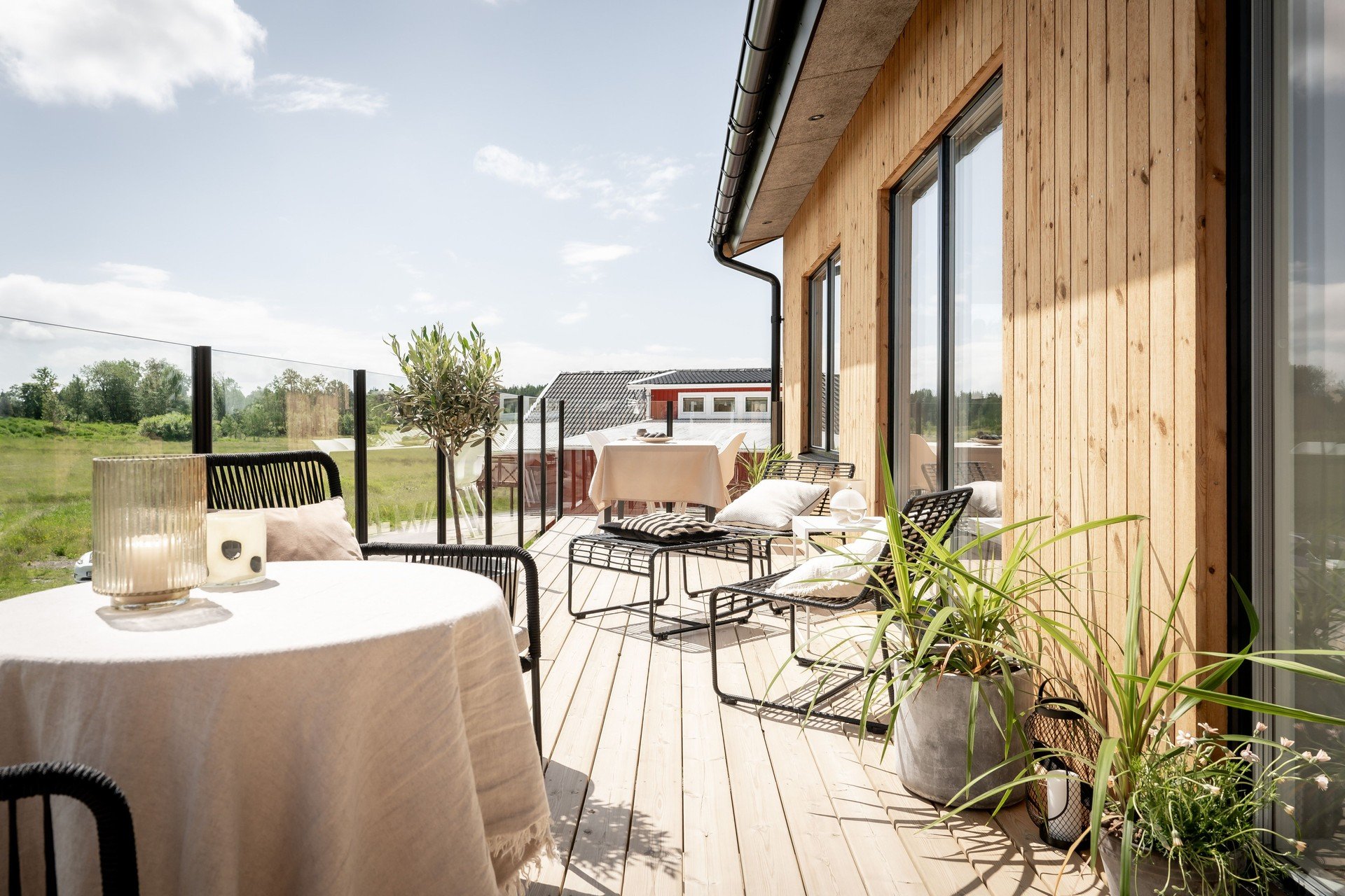 Exteriör träveranda med utemöbler och glasräcke, omgiven av grönska och landskap.