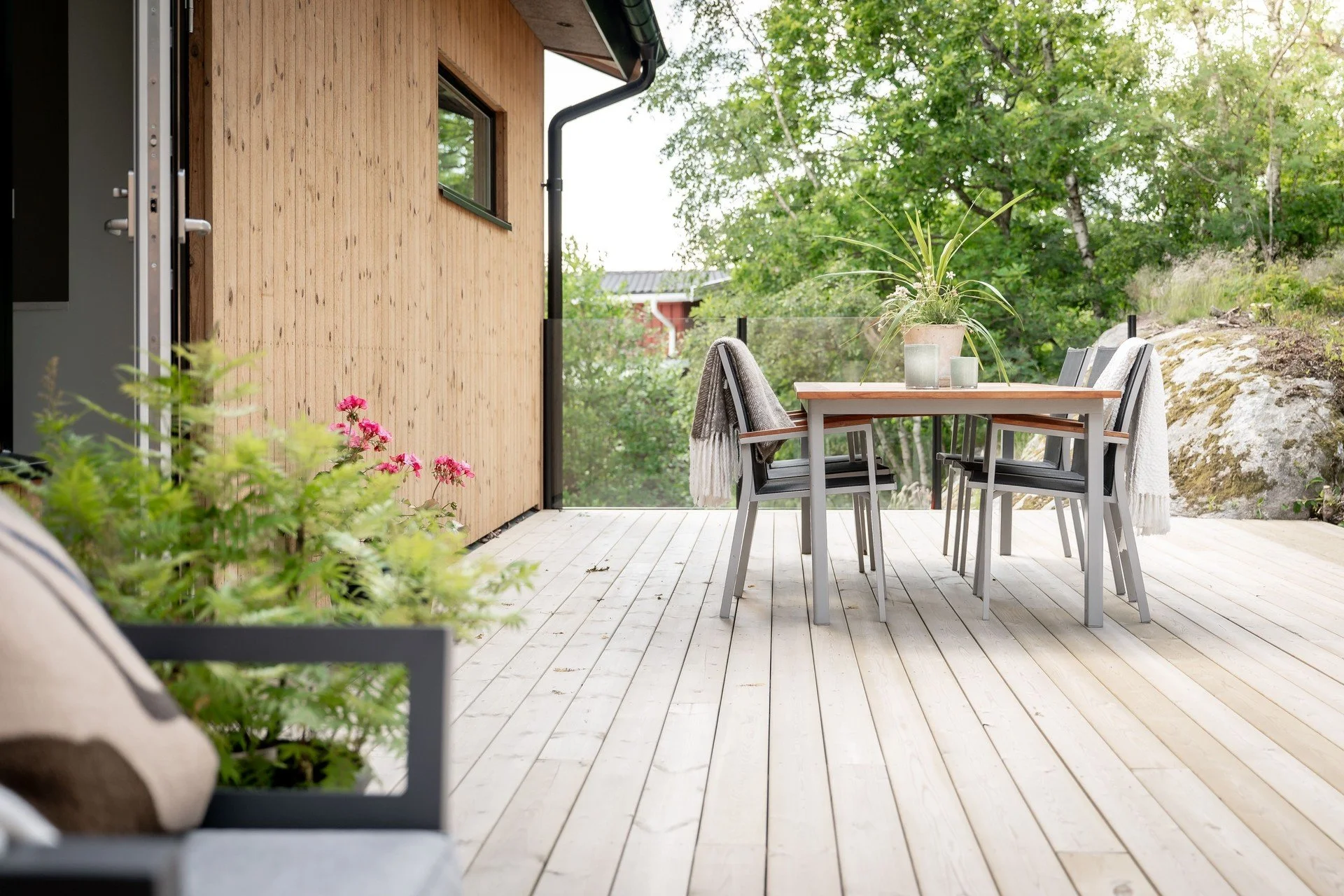 Trädäck med matbord och stolar, krukväxter, filt och grönska i bakgrunden.