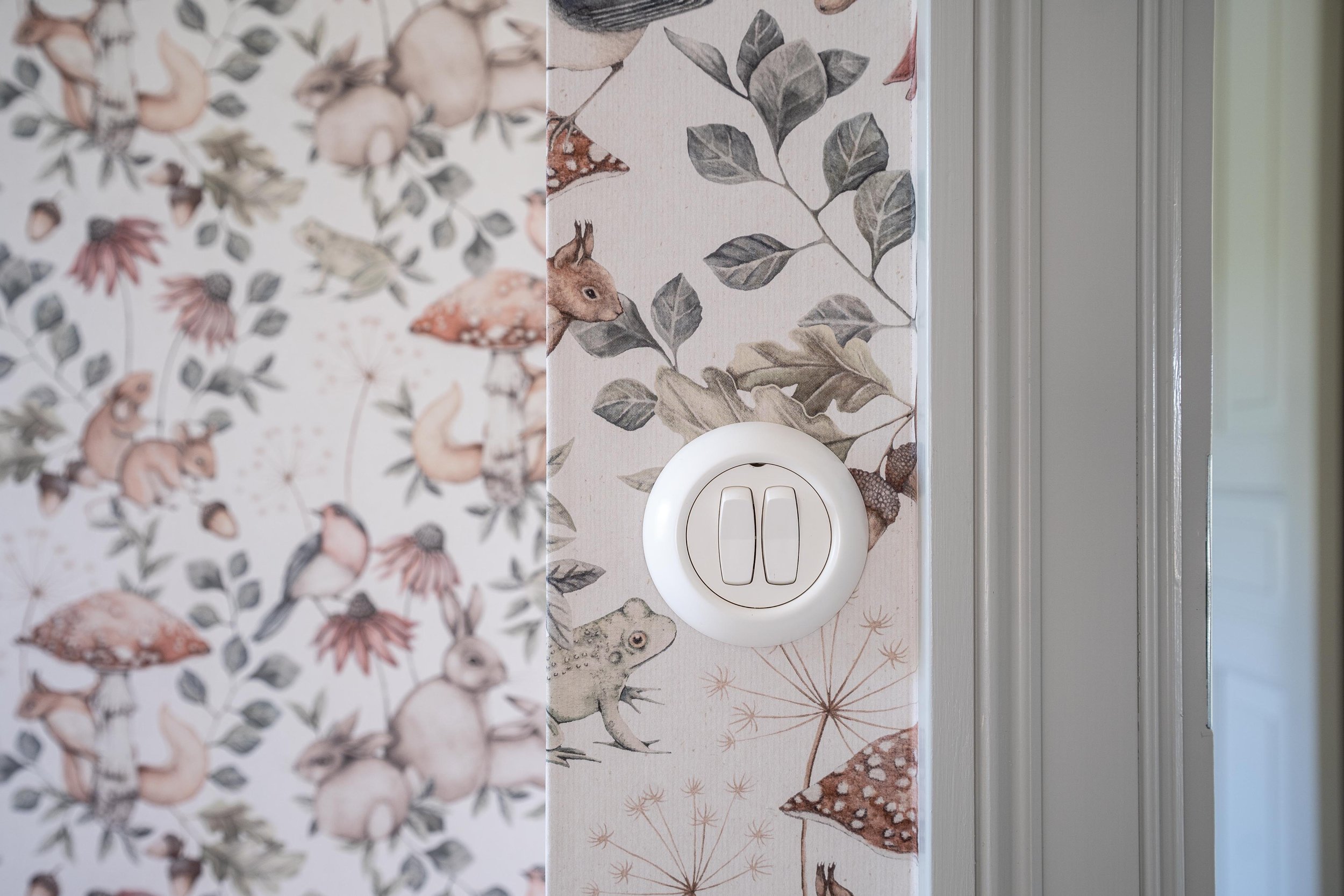 Vägg med barnrumstapet som visar djur och naturmotiv samt en vit strömbrytare. O Lind AB.