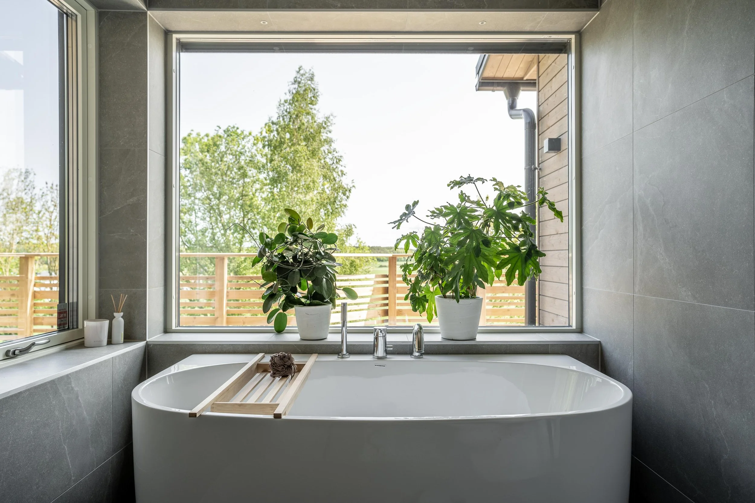 Modernt badrum med stort fönster, badkar och två gröna växter på fönsterbrädan. O Lind AB.