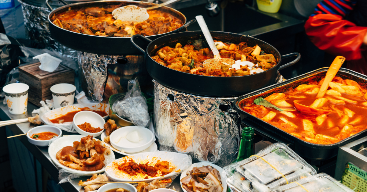 Large pans of tteokbokki and stewed dishes at a Seoul street food market stall.