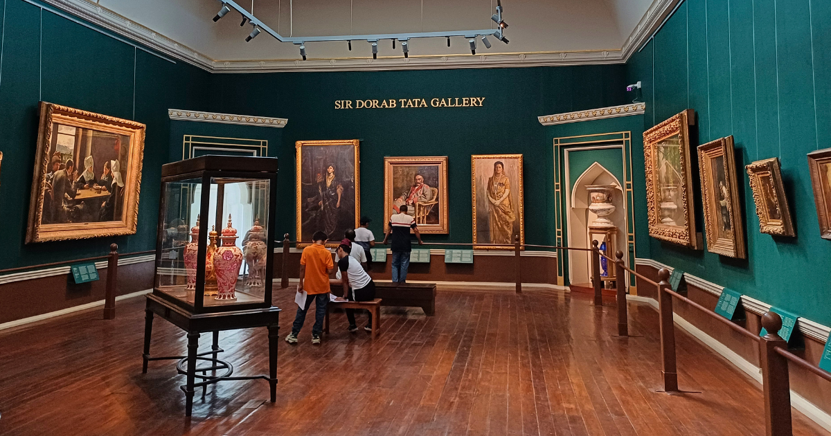 Visitors in the Sir Dorab Tata Gallery at the CSMVS Mumbai, with European oil paintings in gilt frames displayed against dark teal walls and decorative objects in a central glass case.