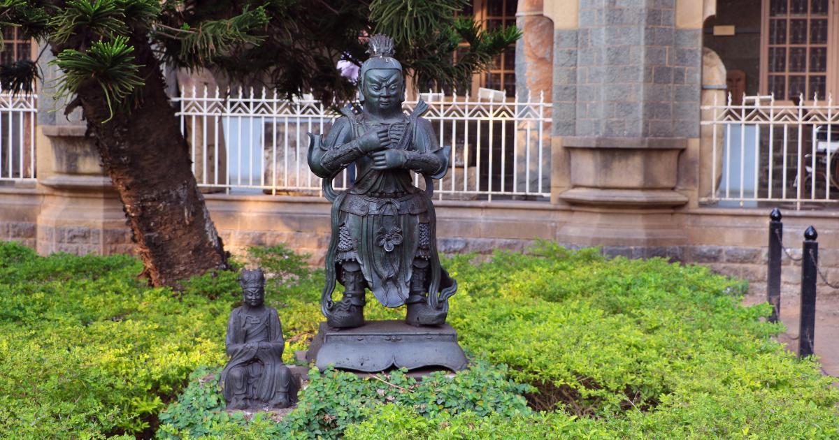 Two bronze East Asian figures displayed in the garden of the CSMVS Mumbai, with the museum's basalt exterior wall and decorative railings visible in the background.