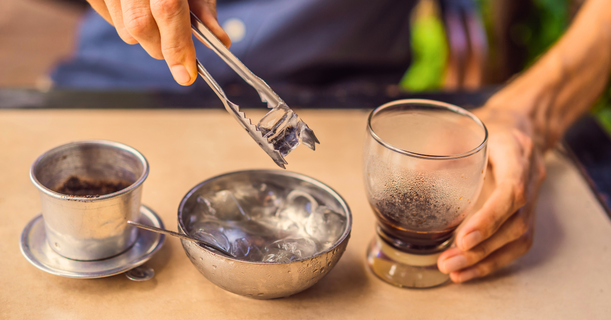 A phin filter on a saucer, a metal bowl of ice and a glass of freshly brewed Vietnamese coffee on a cafe table
