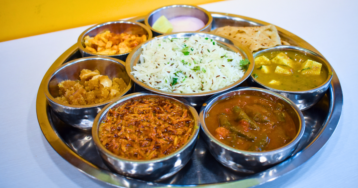 Steel Rajasthani thali with bowls of dal, paneer curry, ker sangri, halwa, jeera rice, papad and pickle