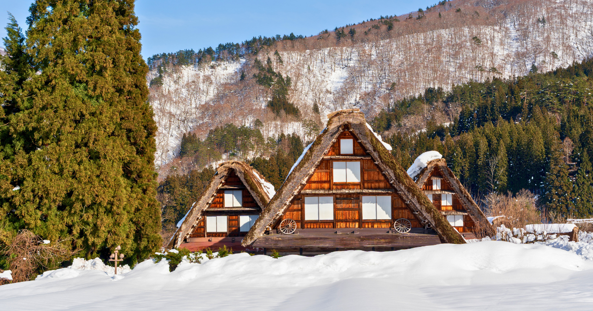 Three traditional thatched roof farmhouses in Shirakawa-go covered in heavy snow with a forested mountain behind