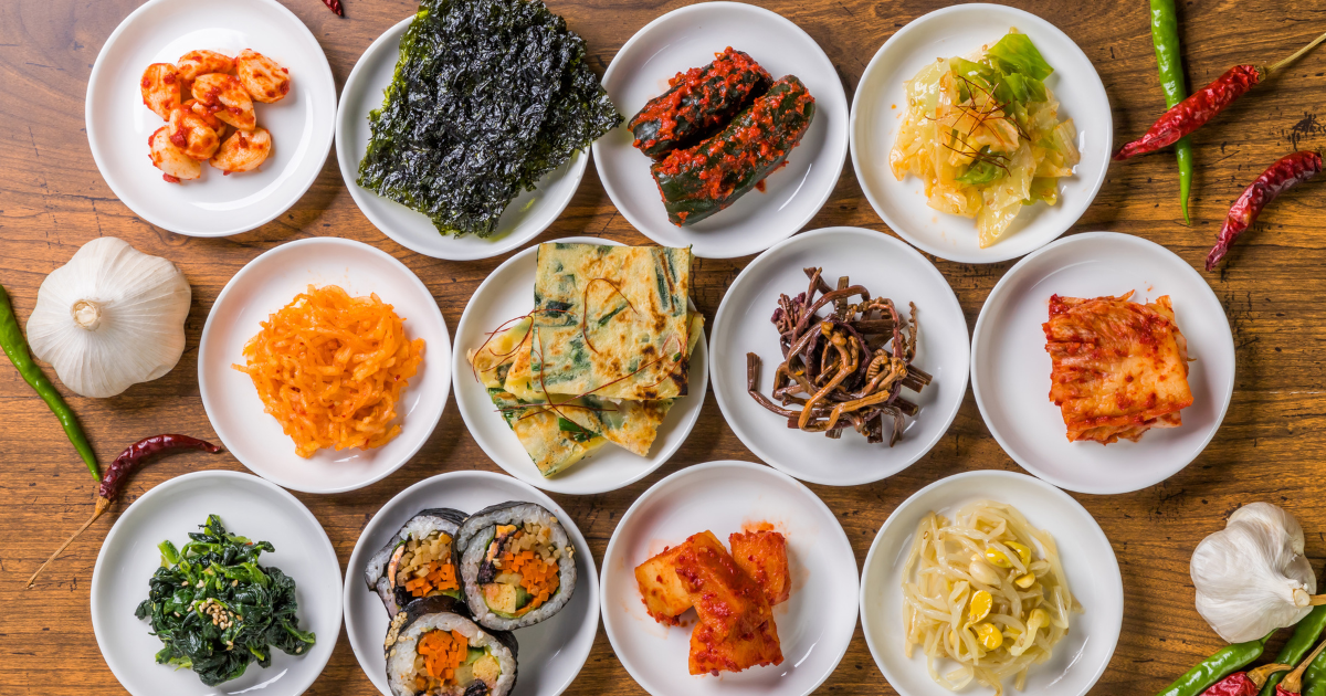 A spread of Korean banchan side dishes including kimchi, gimbap, seasoned spinach, dried seaweed, japchae and pickled radish on small white plates.
