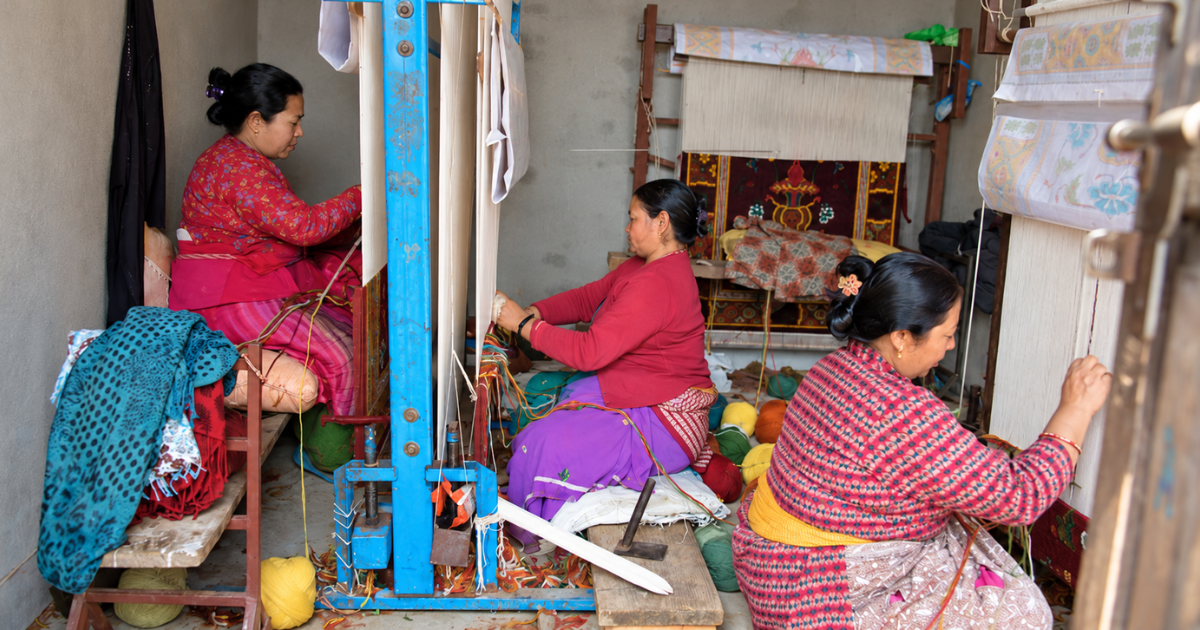 Three women seated at traditional weaving looms, working with coloured yarn to produce patterned carpets.