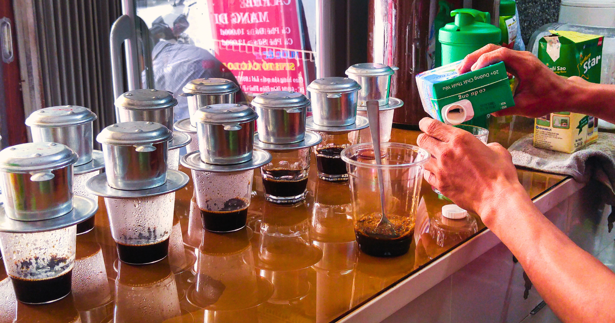 Phin filters dripping coffee into plastic cups at a Vietnamese street stall while condensed milk is poured into a serving cup