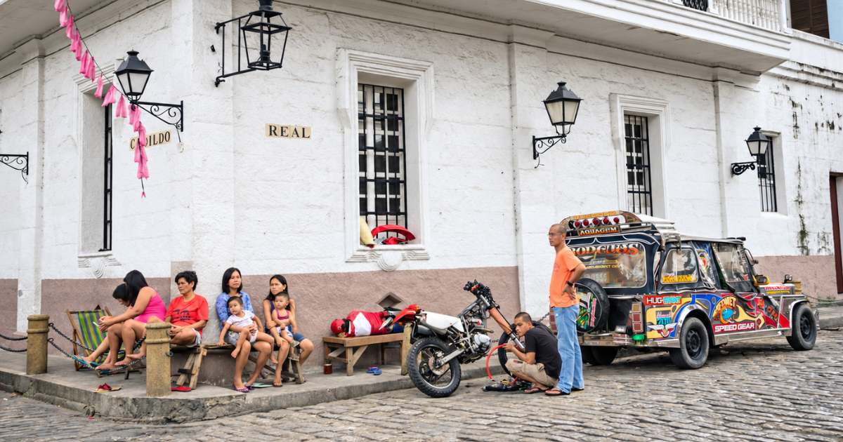 People sitting beside a white colonial-style building at Real and Cabildo streets in Intramuros, with a colourful jeepney and motorcycle nearby.