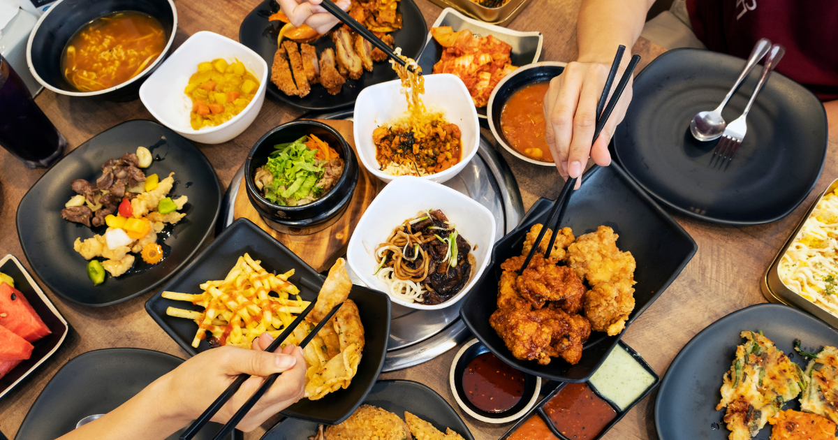 Hands reaching across a crowded table of Korean dishes including fried chicken, noodles, soup, pancakes and dumplings.
