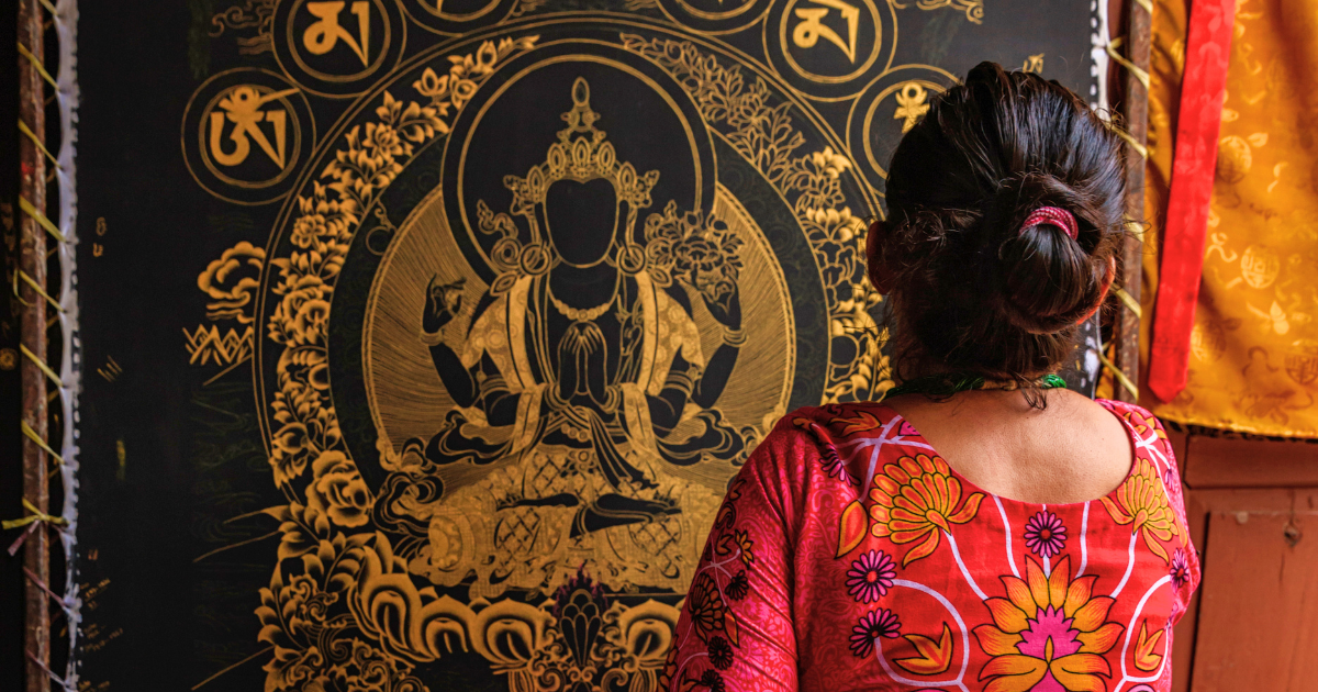 A woman wearing a colourful pink top looking at a black-and-gold Tibetan Buddhist painting depicting a seated religious figure.