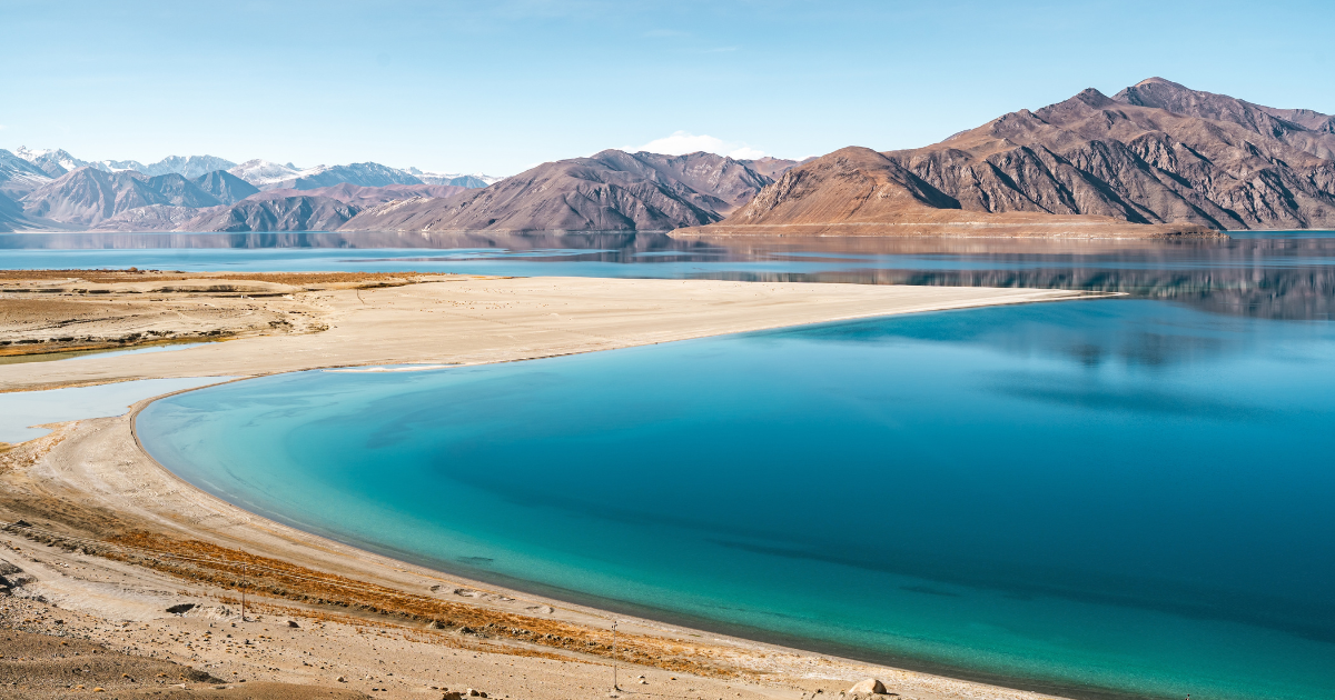 Pangong Lake in Ladakh with turquoise water curving along a sandy shore and barren brown mountains behind.