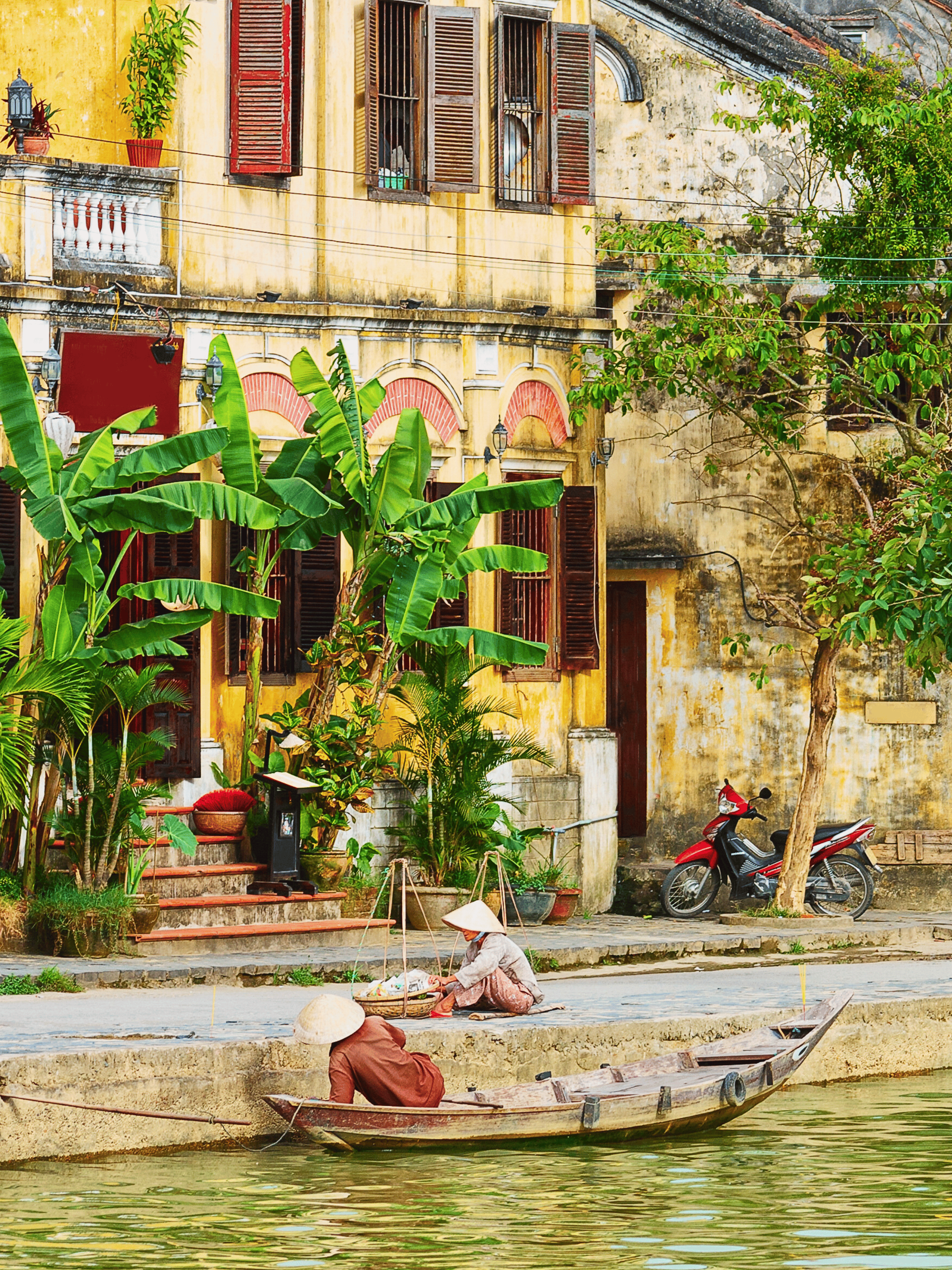 Women in traditional Vietnamese conical hats selling produce on a riverbank near an old building with lush green plants, banana trees, a parked motorbike, and boat in the foreground.