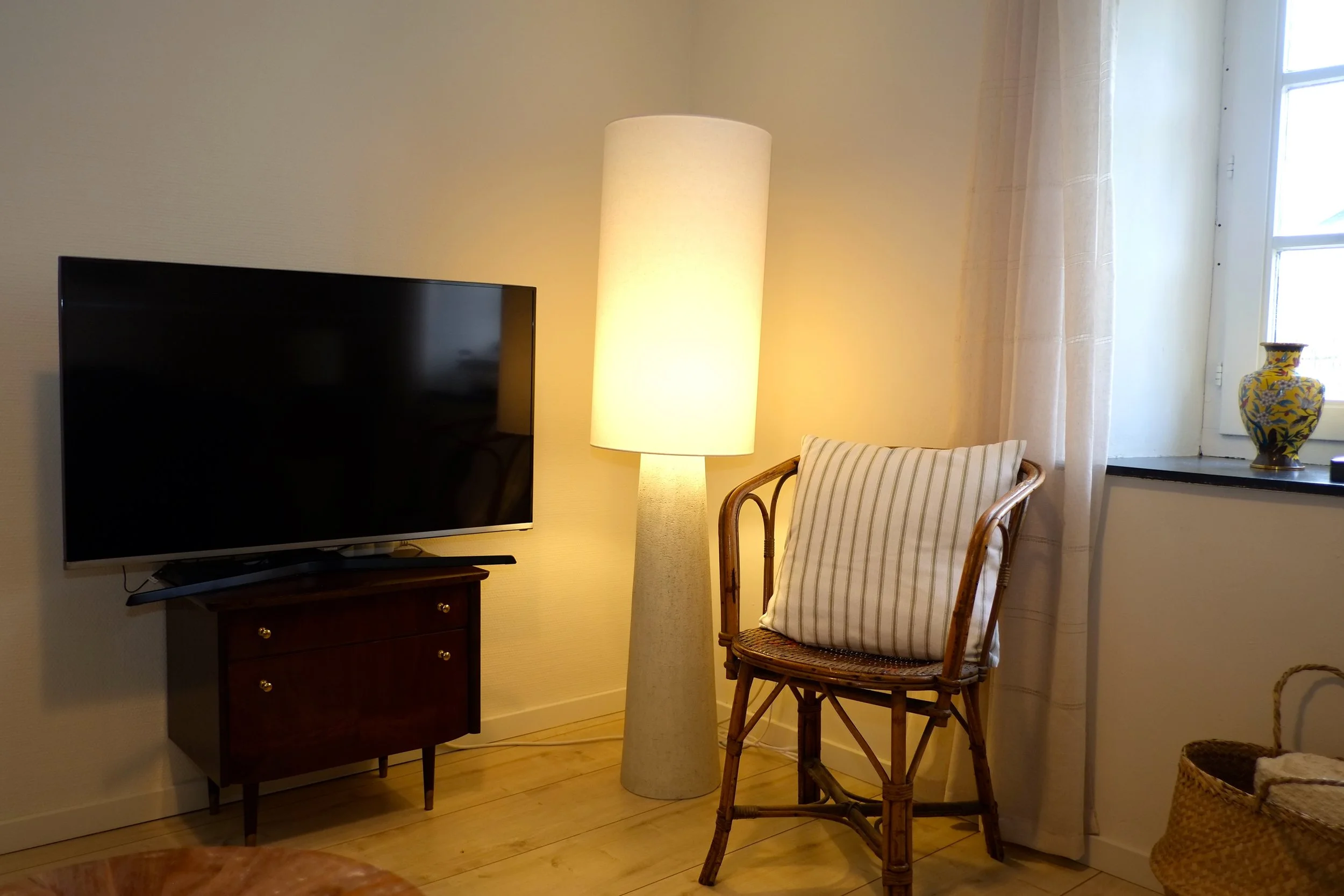 Salon avec télévision, lampe sur pied, chaise en rotin avec coussin, fenêtre avec rideaux et vase décoratif sur le rebord.