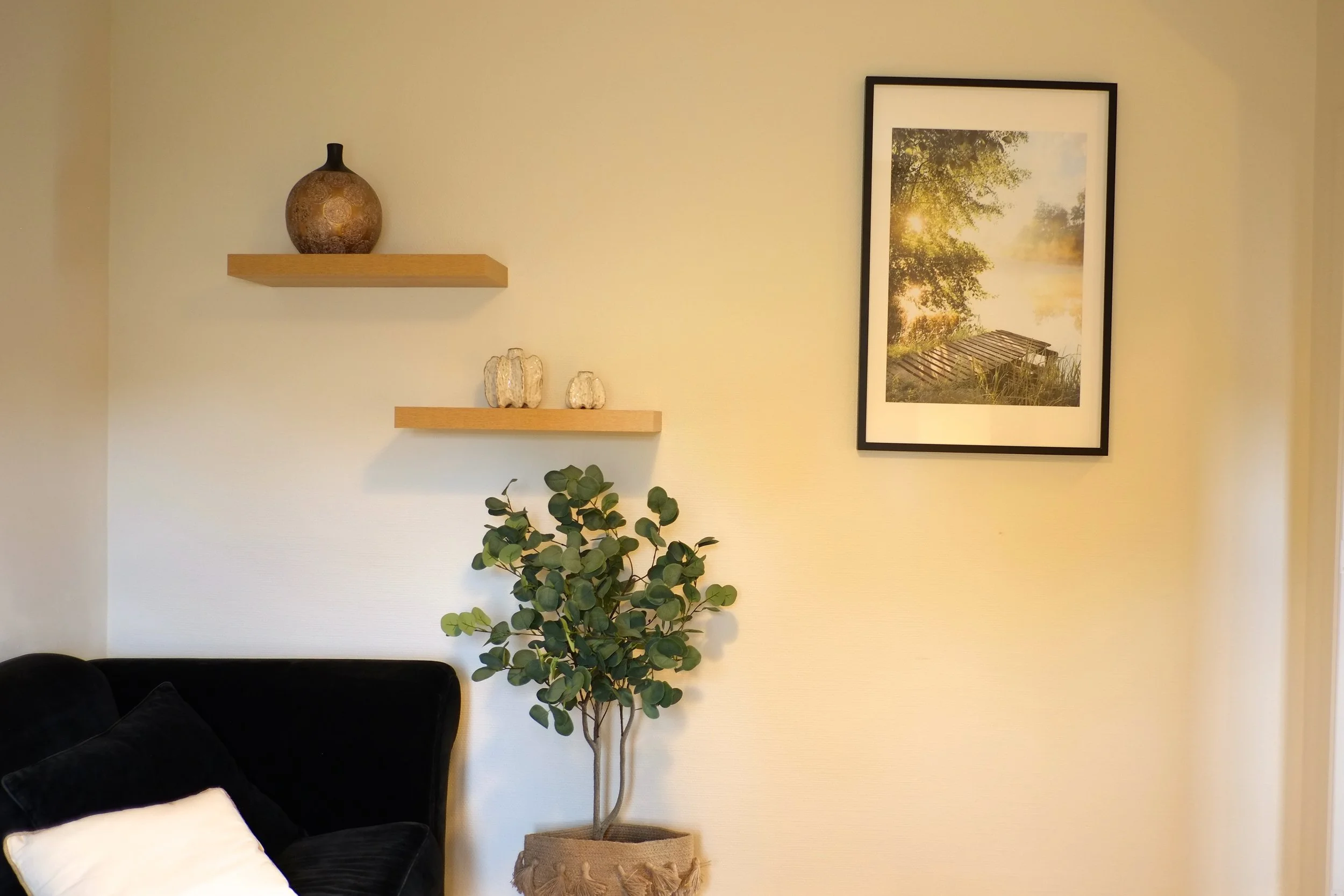 Un mur beige avec deux étagères en bois, une avec une poterie et l'autre avec deux petites décorations, et une photo encadrée d'un paysage avec un pont en bois, une plante verte en pot noir avec un panier en tissu.