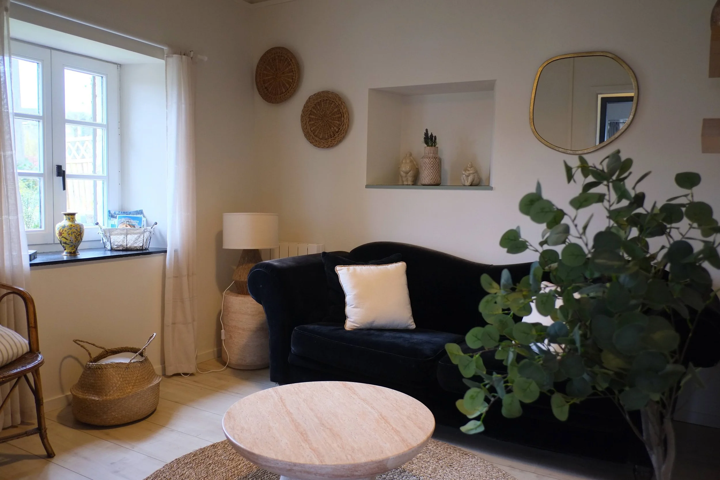 Un salon confortable avec un canapé noir, une table basse en bois, une plante verte, un miroir, une veilleuse, des décorations murales, une fenêtre avec rideaux blancs et une assise en bois avec un coussin à rayures.