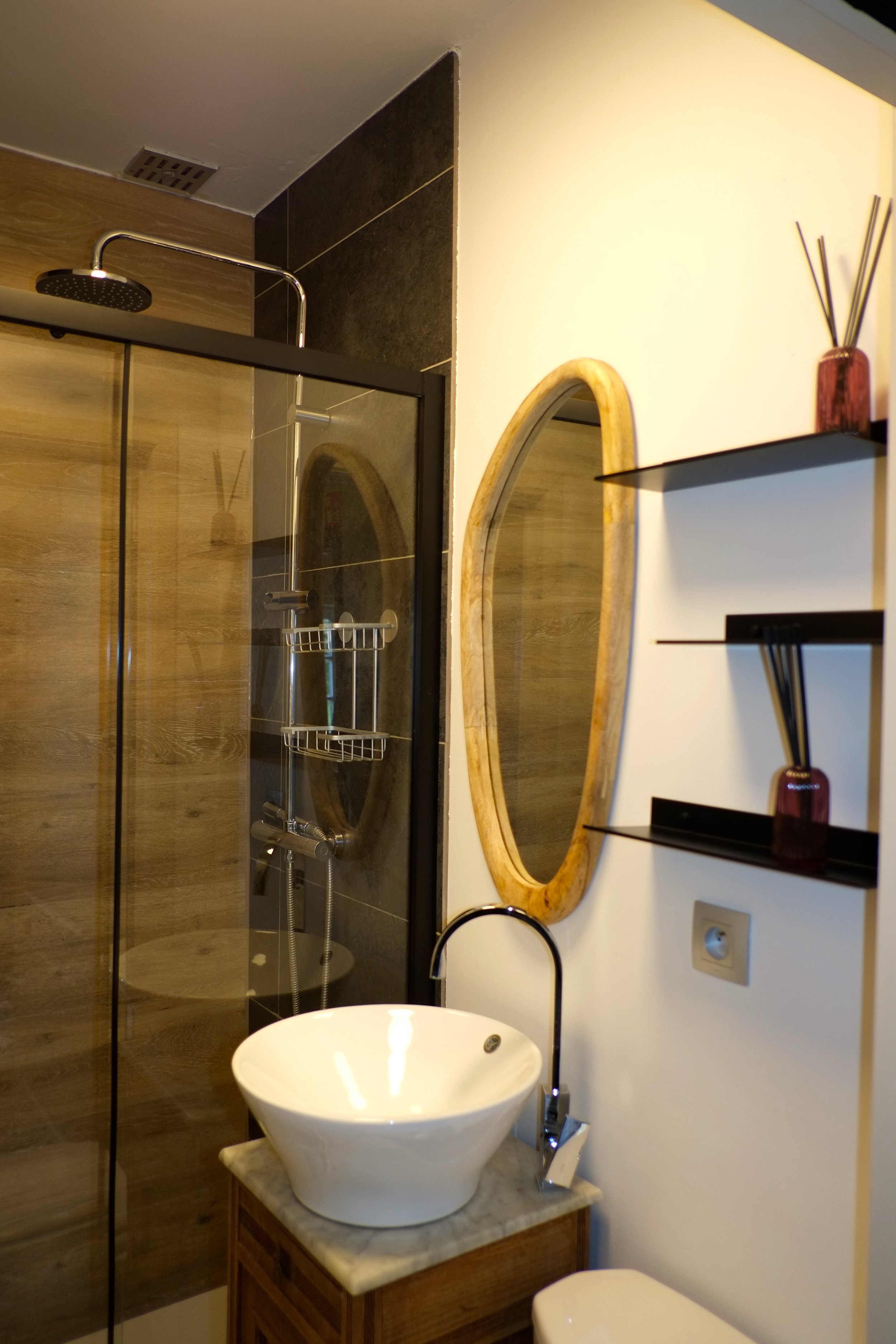 Une salle de bain avec une douche, un lavabo blanc rond sur un meuble en bois, un miroir ovale en bois et une étagère noire avec des décorations en rouge, en forme de vase avec des tiges.