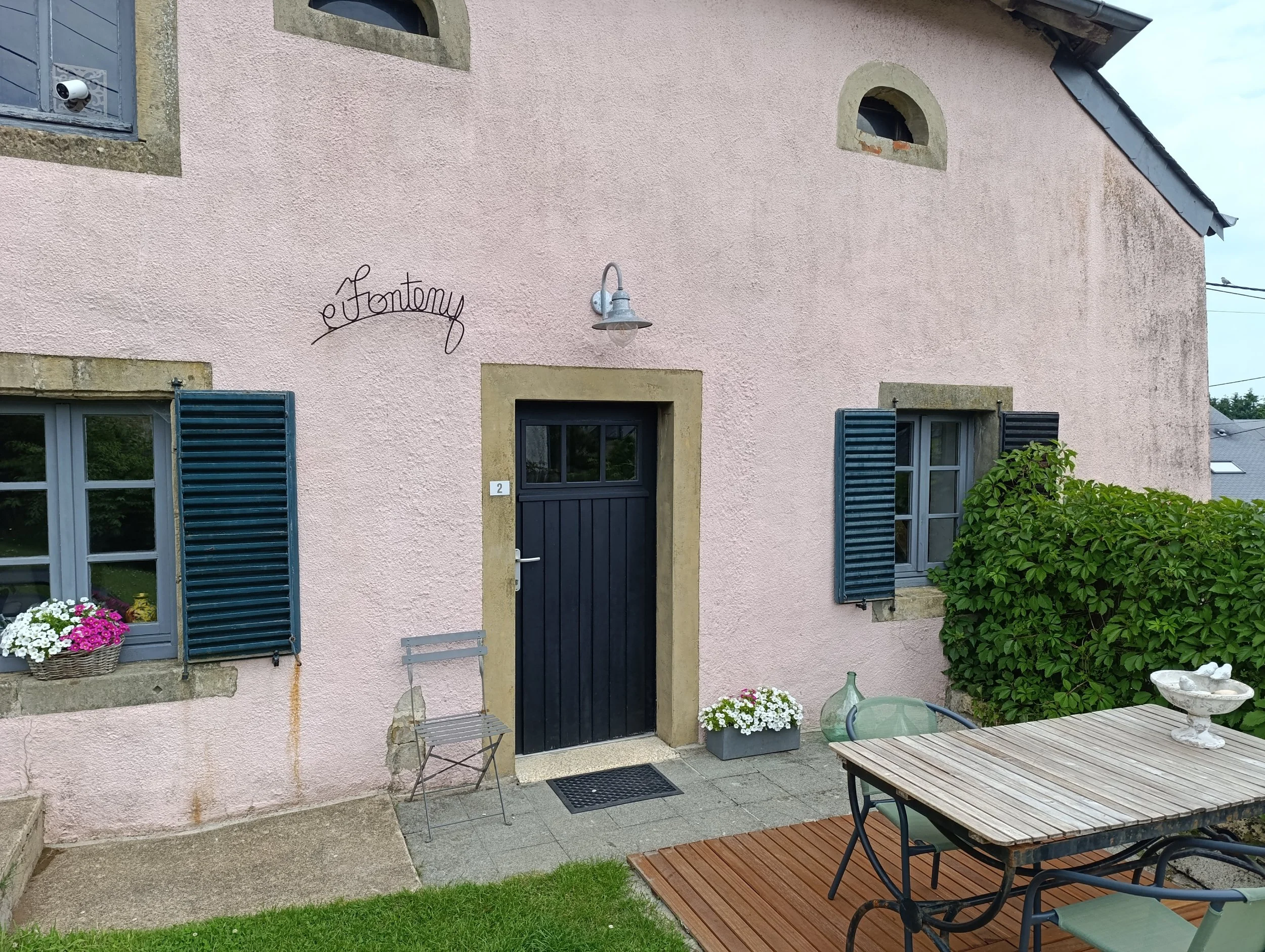 Une maison avec des murs roses, des volets bleus et une porte noire. Il y a une table en bois avec des chaises et une petite cour avec des fleurs et des plantes. On voit également une enseigne en fil de fer avec le nom 'e Fontony'.