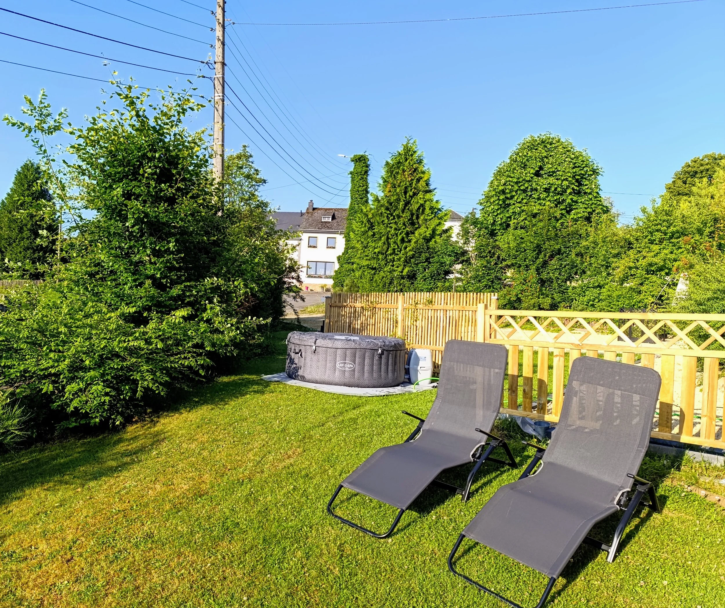 Jardin avec deux chaises longues, piscine gonflable, pelouse verte, arbres et haies, ciel bleu.