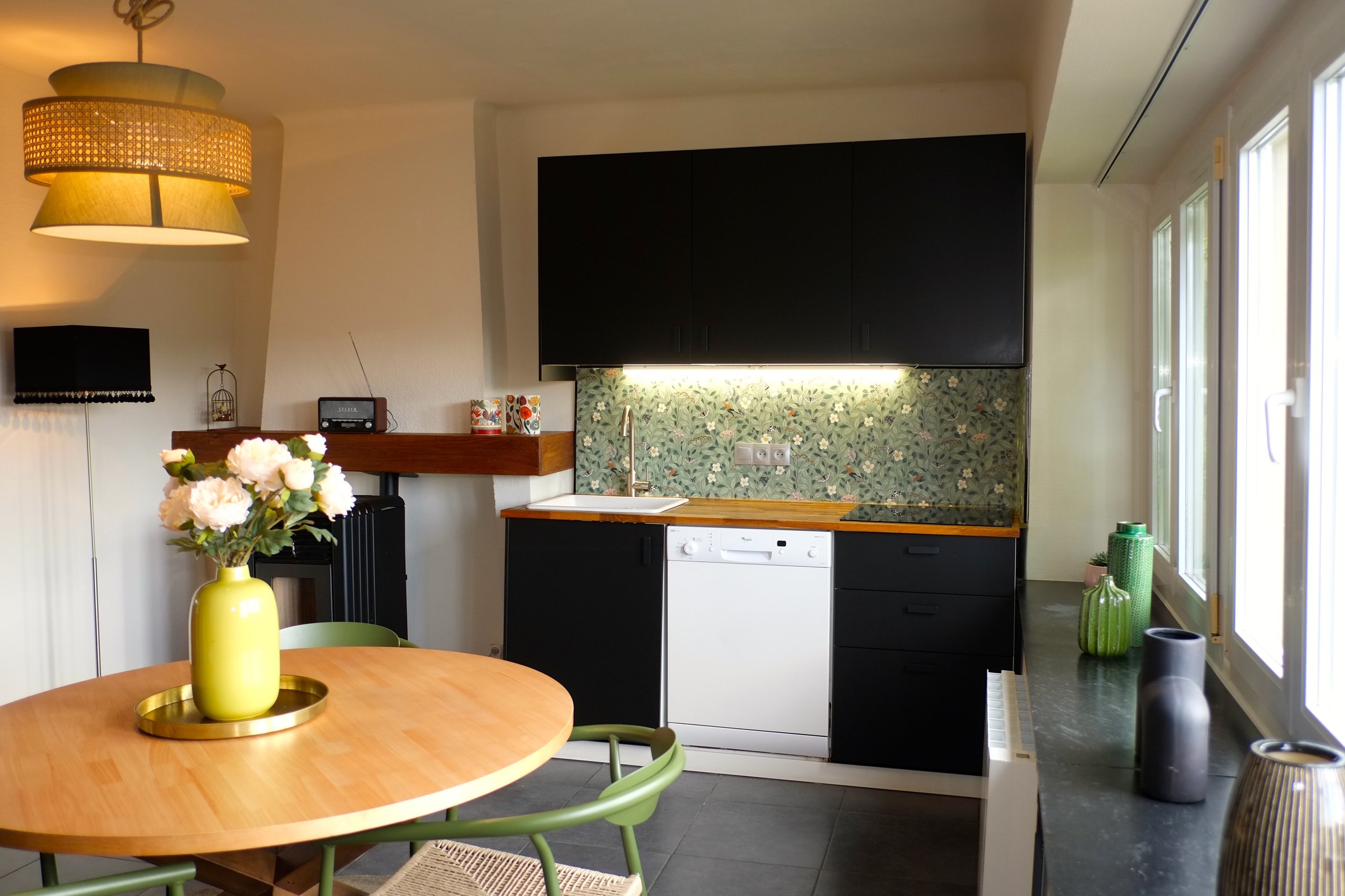 Cuisine avec mobilier noir, plans de travail en bois, électroménagers, décoration florale et luminaires modernes.