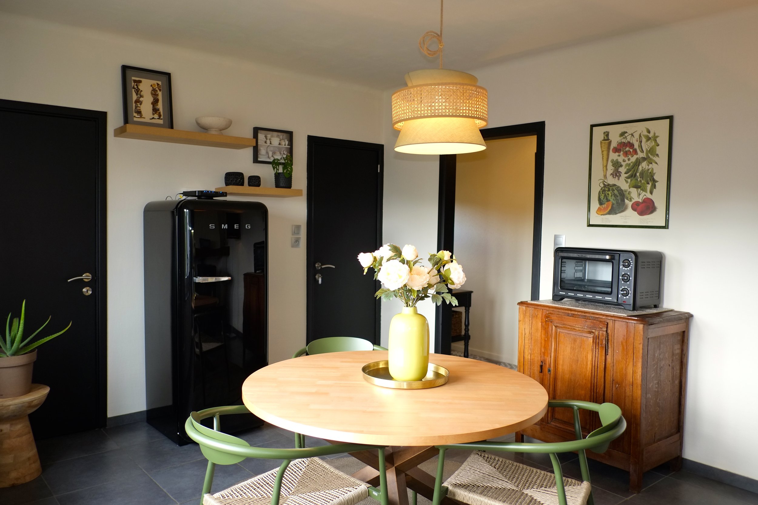 Une salle à manger avec un meuble en bois, un micro-ondes, un tableau avec des fruits, une grande lampe suspendue, une table ronde en bois avec un vase de fleurs, quatre chaises vertes en métal avec sièges en paille, un réfrigérateur noir, des étagères avec décorations, un porte-clés et un mur blanc.