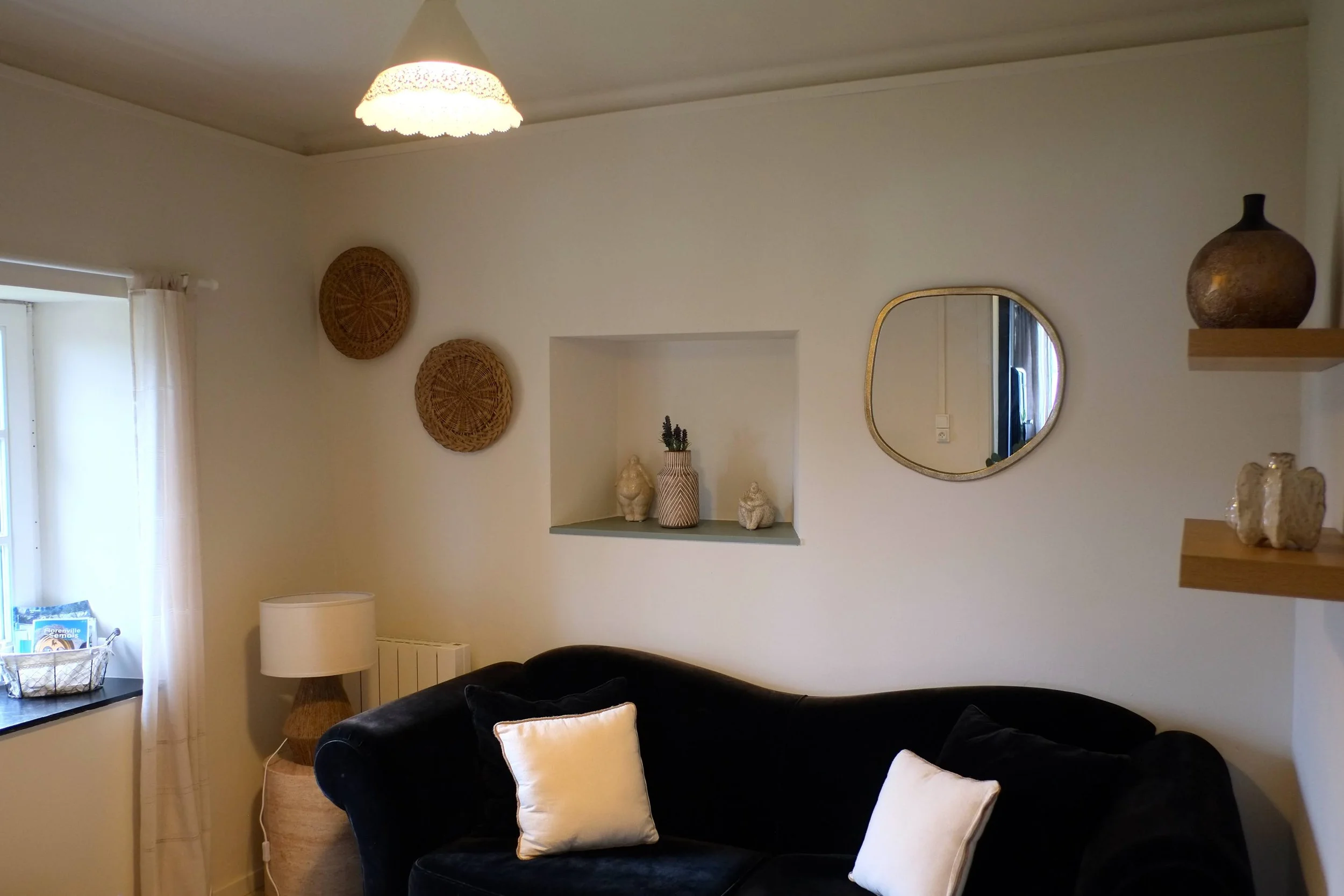 Salon avec un canapé noir, deux coussins blancs, une lampe, une fenêtre avec rideaux blancs, des décorations en bambou et en céramique sur les murs et une étagère en bois avec des vases.