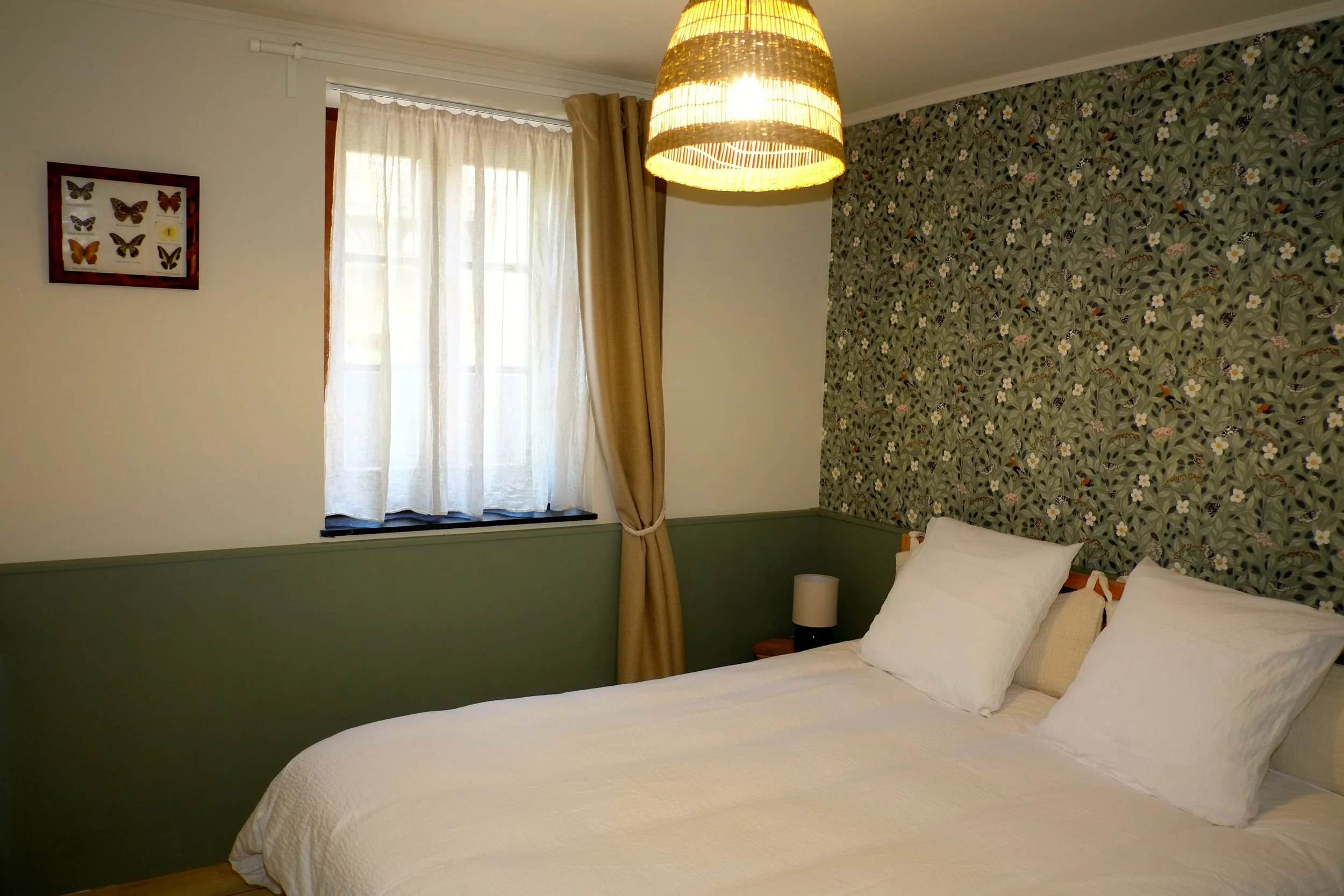 Chambre avec lit double, trois oreillers, mur avec papier peint à motifs floraux, lampe suspendue en rotin, fenêtre avec rideaux blancs et beige, tableau de papillons