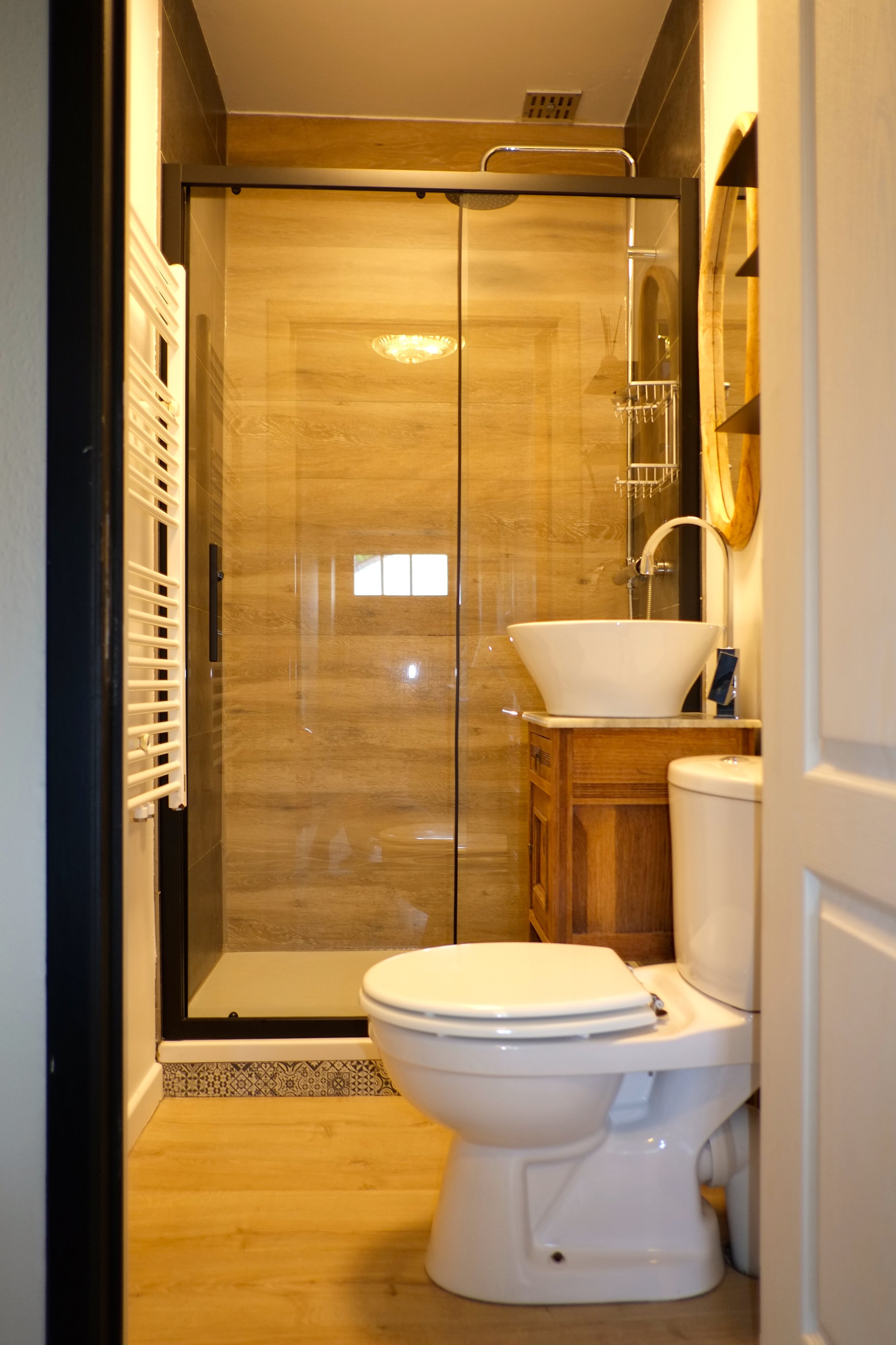 Une petite salle de bain avec une toilette, un lavabo sur un meuble en bois, une douche avec porte en verre, et un miroir rond.