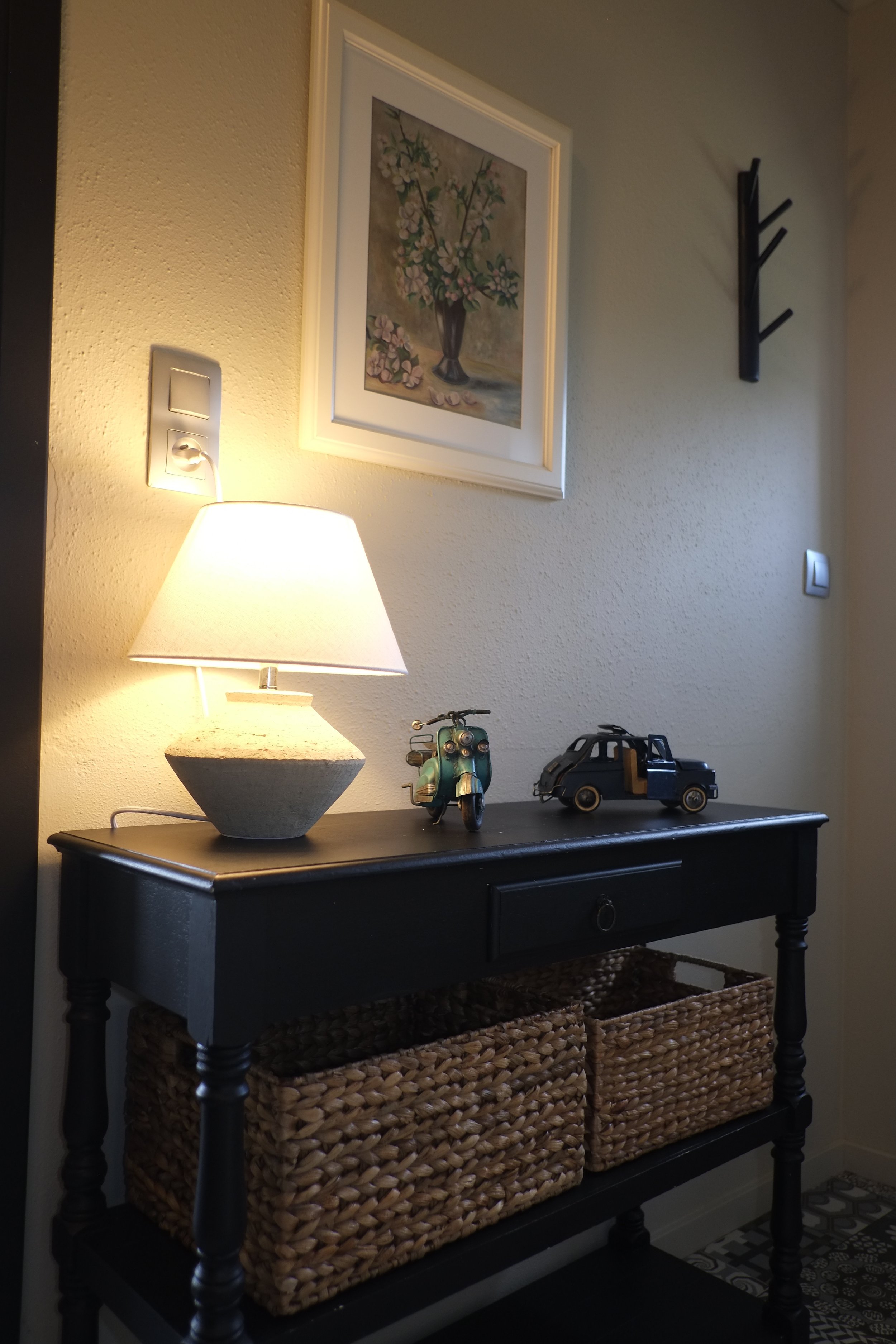 Console noire avec deux paniers en osier en dessous, lampe de table, figurines de scooter et voiture, tableau avec fleurs au mur, porte-manteau en bois noir, interrupteur