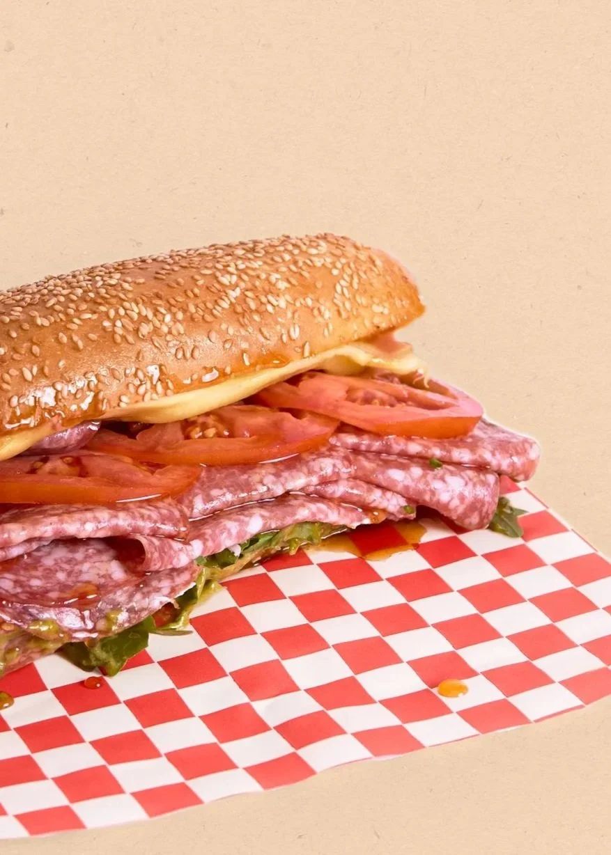 A hoagie sandwich with sliced tomatoes, lettuce, and multiple types of deli meat on a sesame seed hoagie bun, placed on red and white checkered paper.