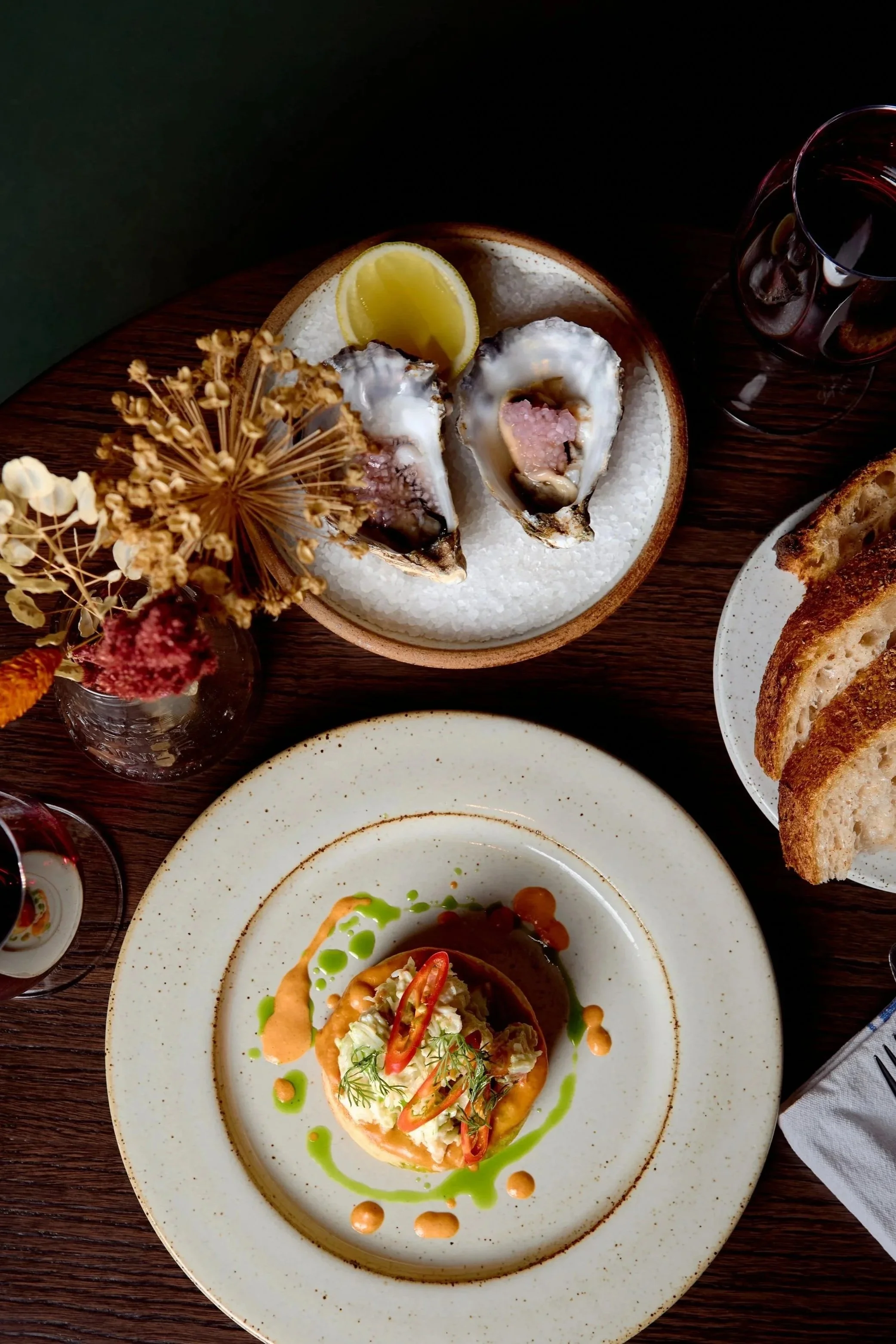 Spouse Studio. Gemma Tree. A top-down view of a meal with oysters on a bed of ice, lemon wedge, glass of red wine, toasted bread slices, and a plated dish with a colorful vegan or vegetarian meal garnished with herbs, chili slices, and sauces.