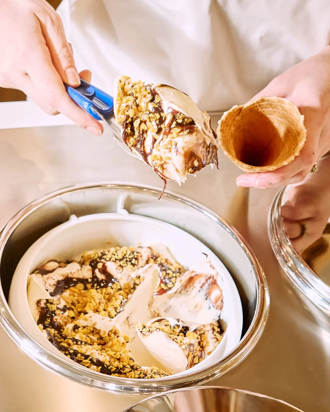 Spouse Studio. Gemma Tree. Person scooping ice cream topped with chocolate sauce and crushed nuts into a waffle cone.