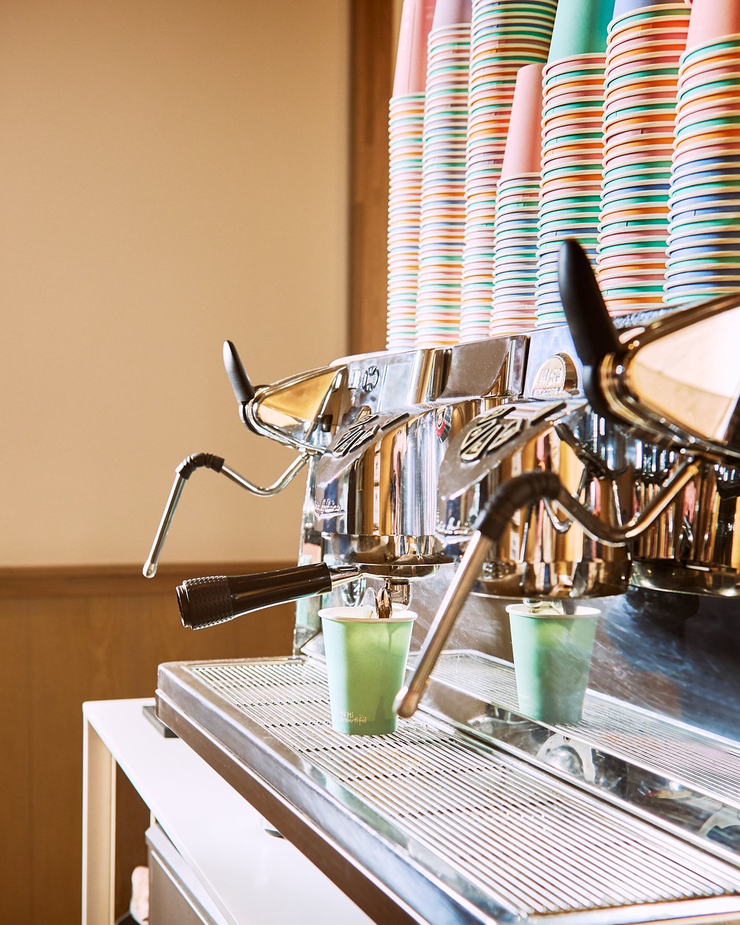 Spouse Studio. Gemma Tree. A stainless steel espresso machine pouring coffee into a green paper cup, with colorful striped paper cups stacked behind it.