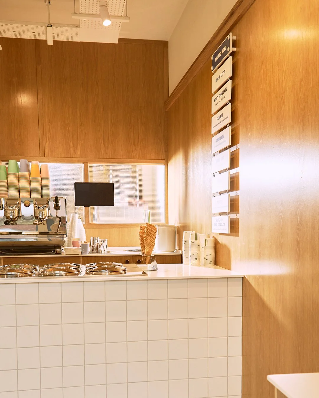 Spouse Studio. Gemma Tree. Coffee shop counter with coffee machine, paper cups, ice cream cone, and menu board on the wooden wall.