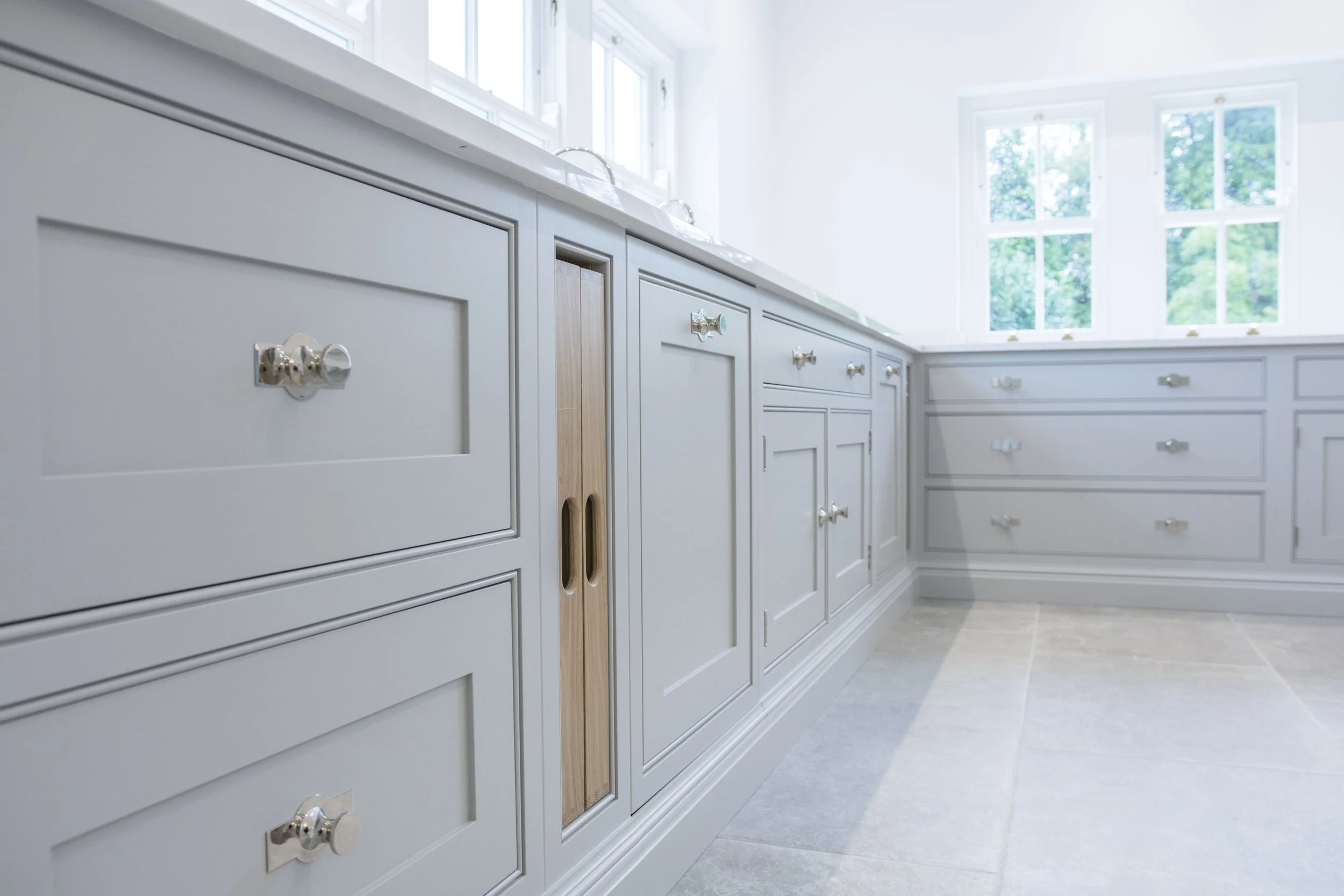 Light grey kitchen cabinets with silver knobs, set against a bright, airy room with large windows and a tiled floor.