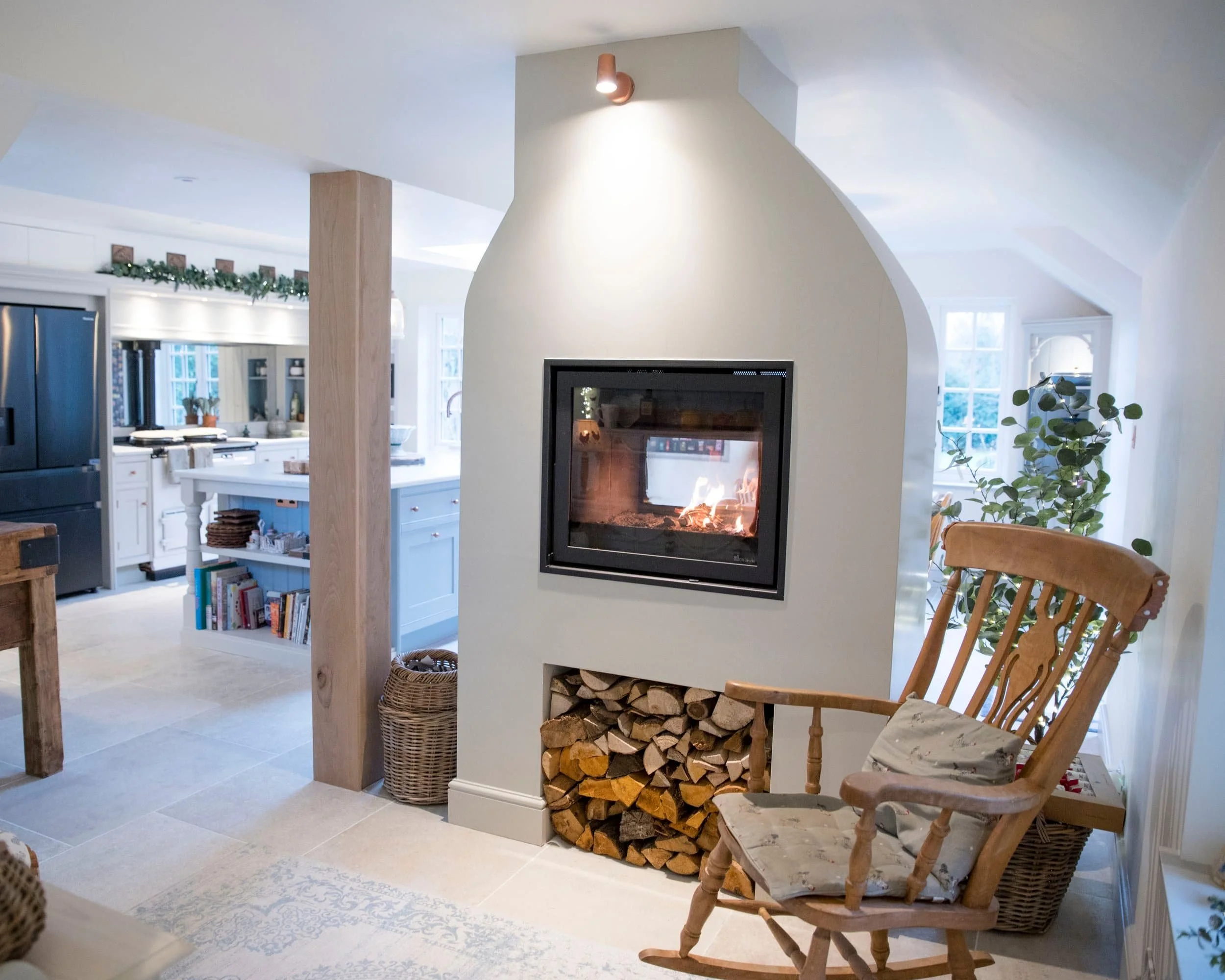 Cozy living room with a modern fireplace, stacked firewood, a wooden rocking chair, and a bright, airy kitchen in the background.