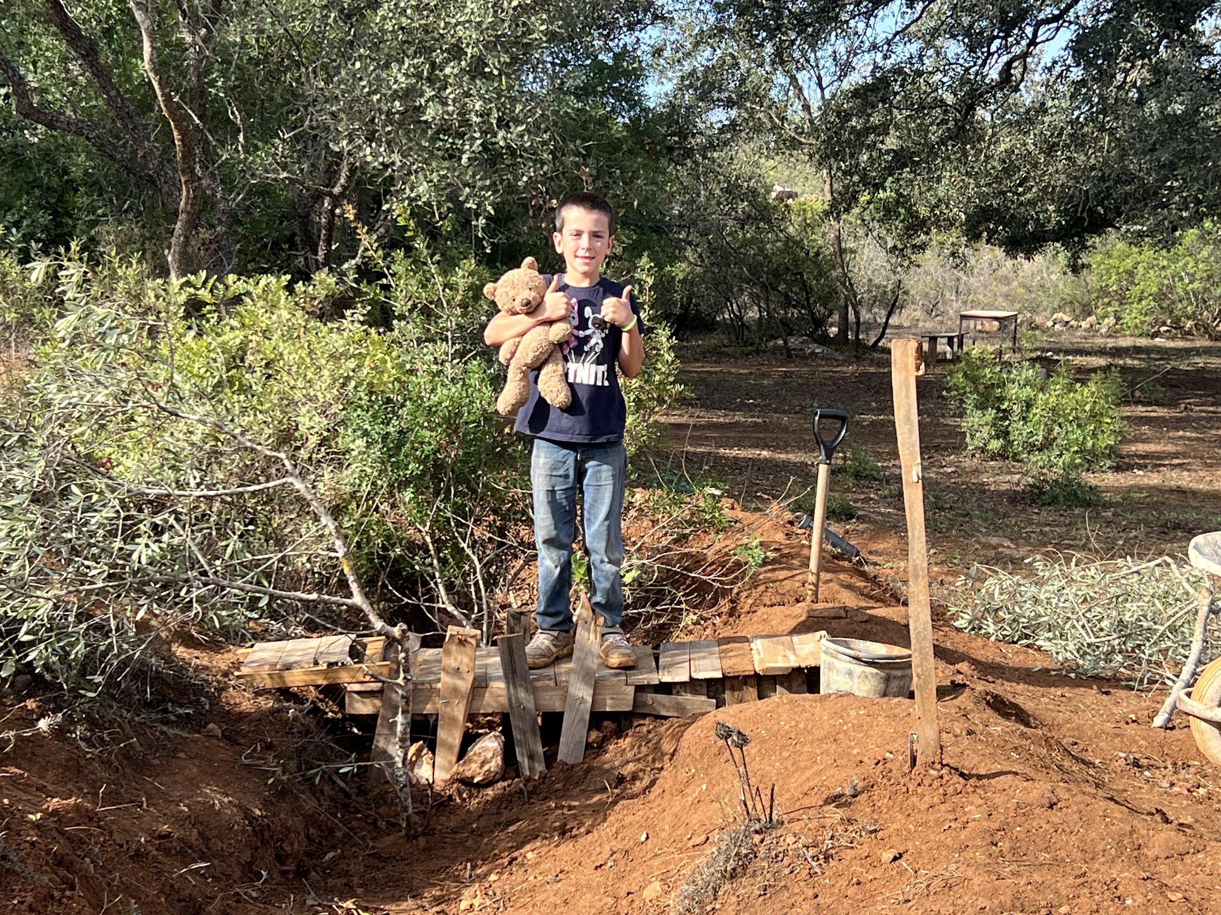 Young boy with teddy bear standing on a wooden bridge in a garden with surrounding trees and bushes.