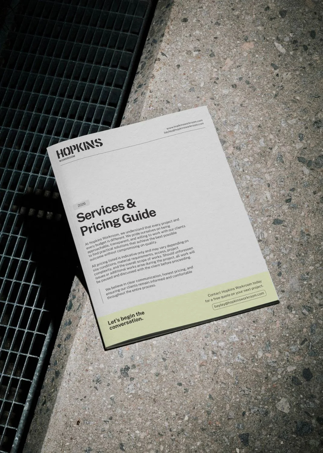 A printed Services & Pricing Guide for Hopkins Workroom placed on a speckled concrete floor, near a metal grate.